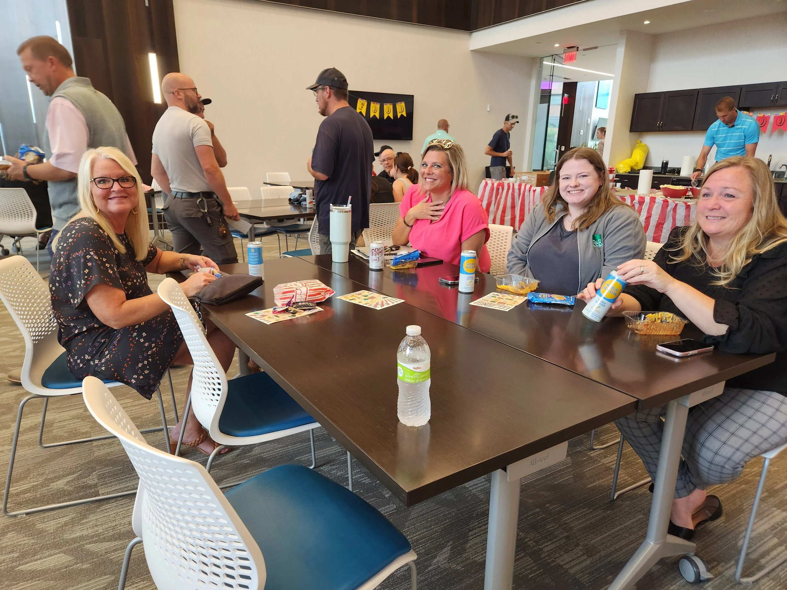 Four women sitting at a table with snacks and drinks, smiling and interacting in a casual indoor setting with people in the background.