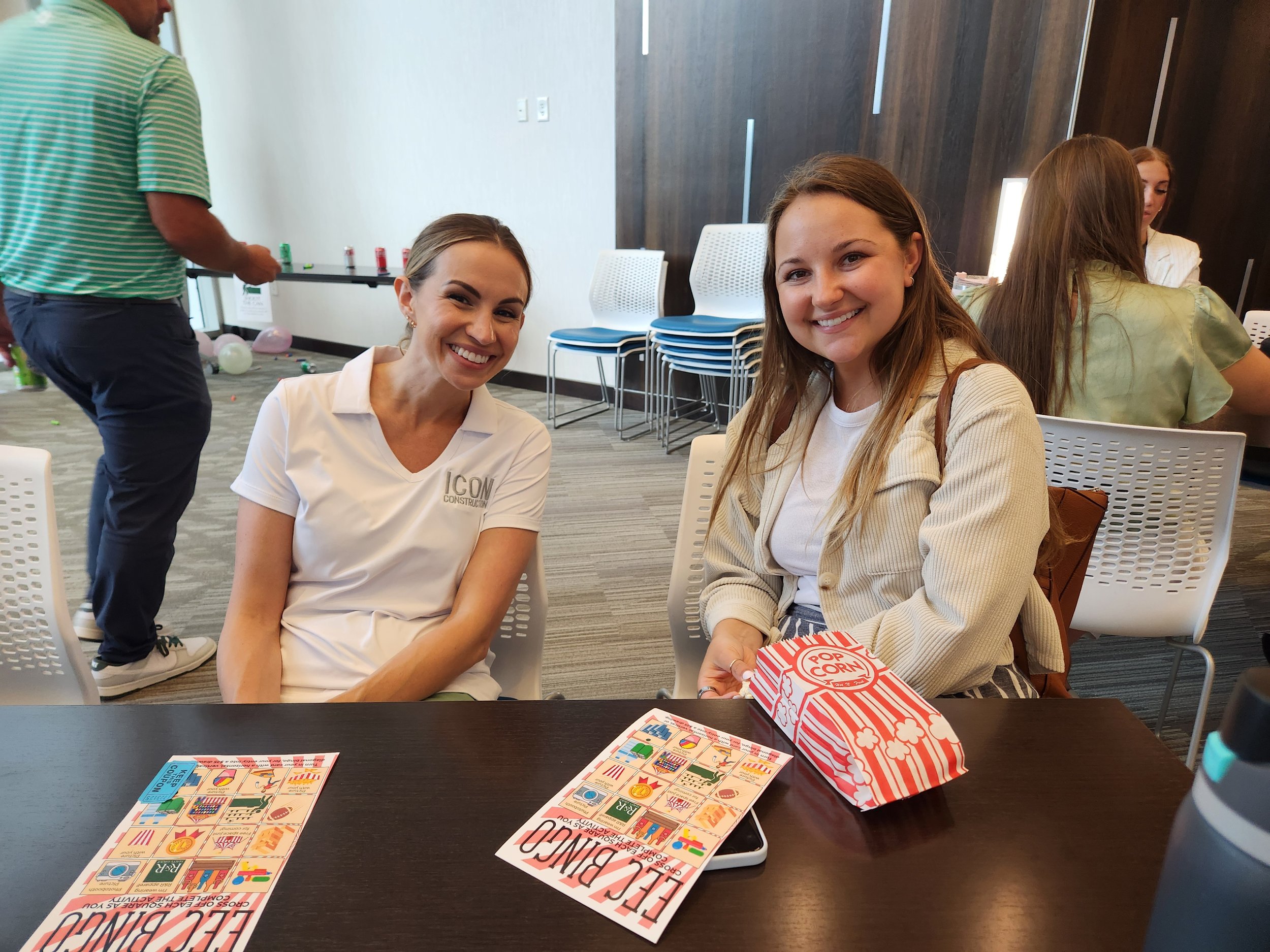 Two women sitting at a table, smiling. One is wearing a white shirt with 'ICON' on it, and the other has a popcorn box and a game sheet in front of her. There are other people and chairs in the background.