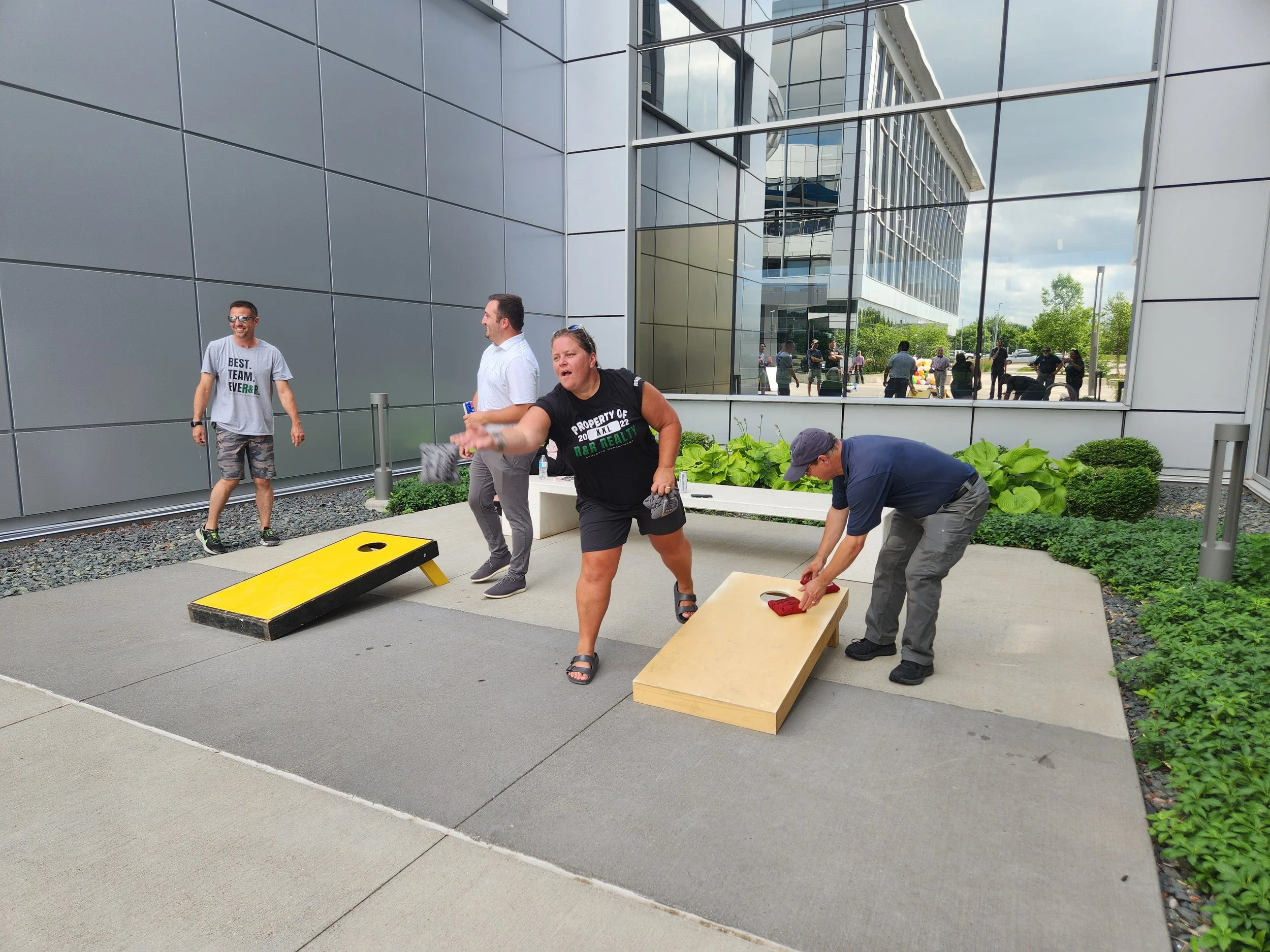 Four people playing cornhole outside a modern building. Two are actively tossing bean bags into holes on game boards, one is watching, and another is walking past. The background shows a reflective glass building and people outside.