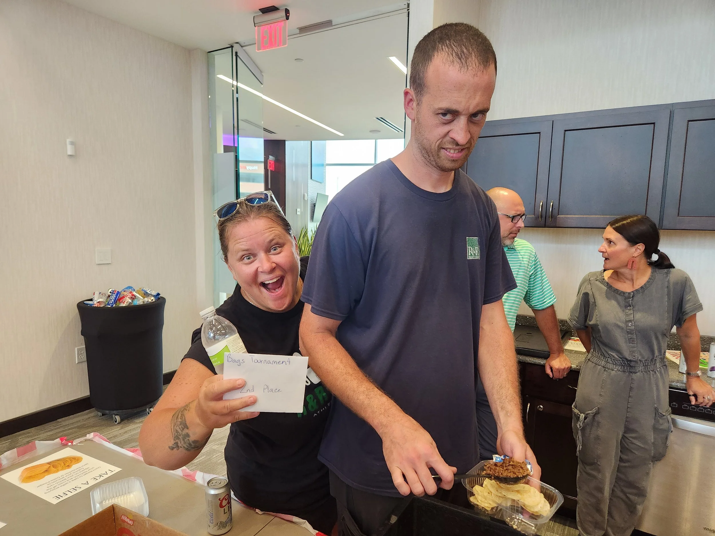 A woman holding a bottle and a sign that reads 'Bags Tournament 2nd Place' smiling and posing behind a man in a blue t-shirt who is serving food on a tray in an indoor setting. Two other people are in the background near kitchen cabinets.