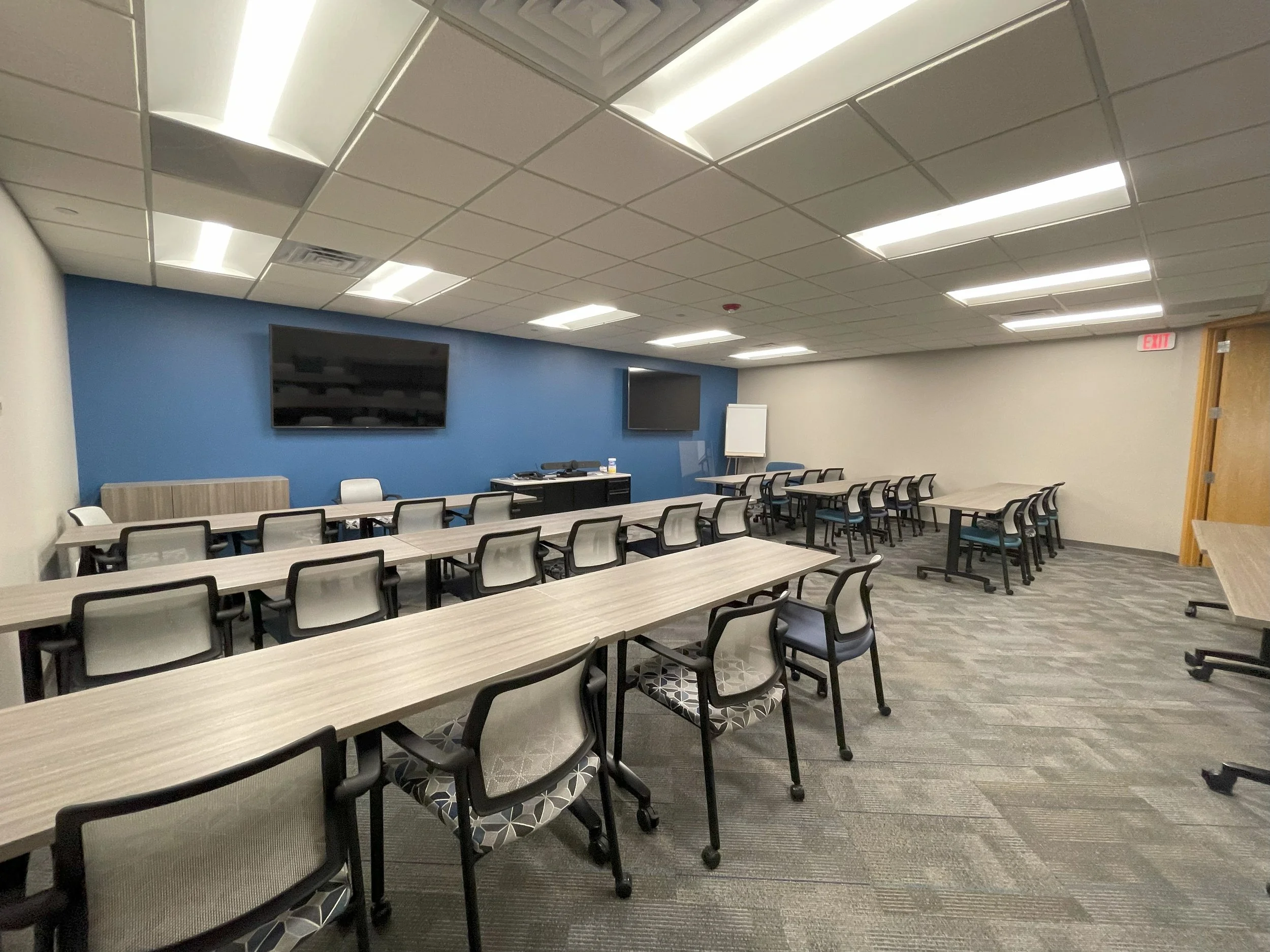 Empty conference room with long tables, chairs, two large screens on blue wall, whiteboard, and exit door.