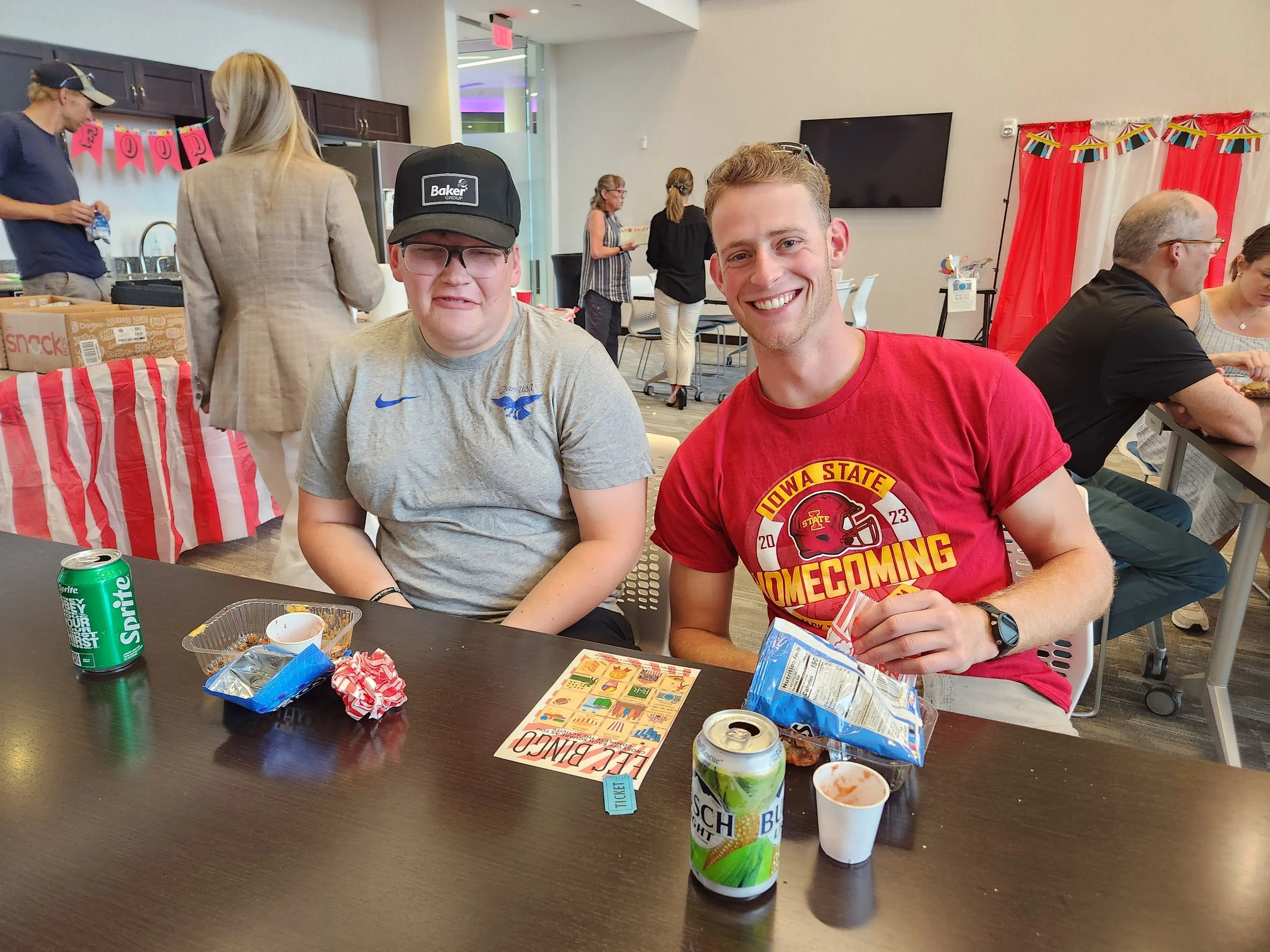 Two young men sitting at a table during a gathering or event, with snacks and drinks in front of them. One wears a gray t-shirt and glasses, the other wears a red Iowa State homecoming t-shirt, smiling at the camera. In the background, there are othe