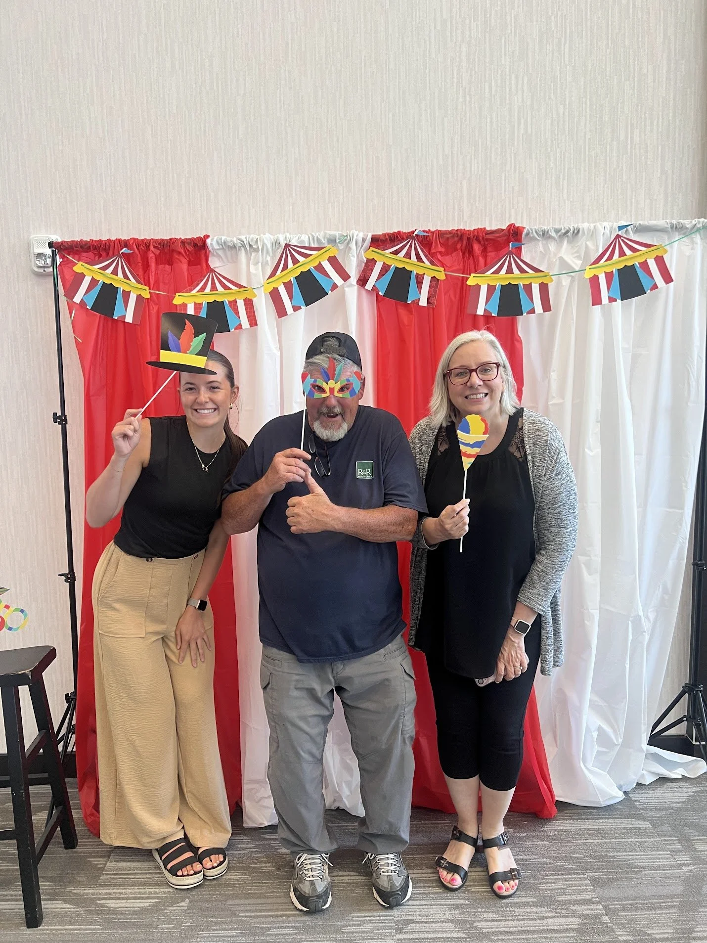 Three people posing in front of a red and white circus-themed backdrop, holding paper props like a hat and a mask, smiling and enjoying a festive event.