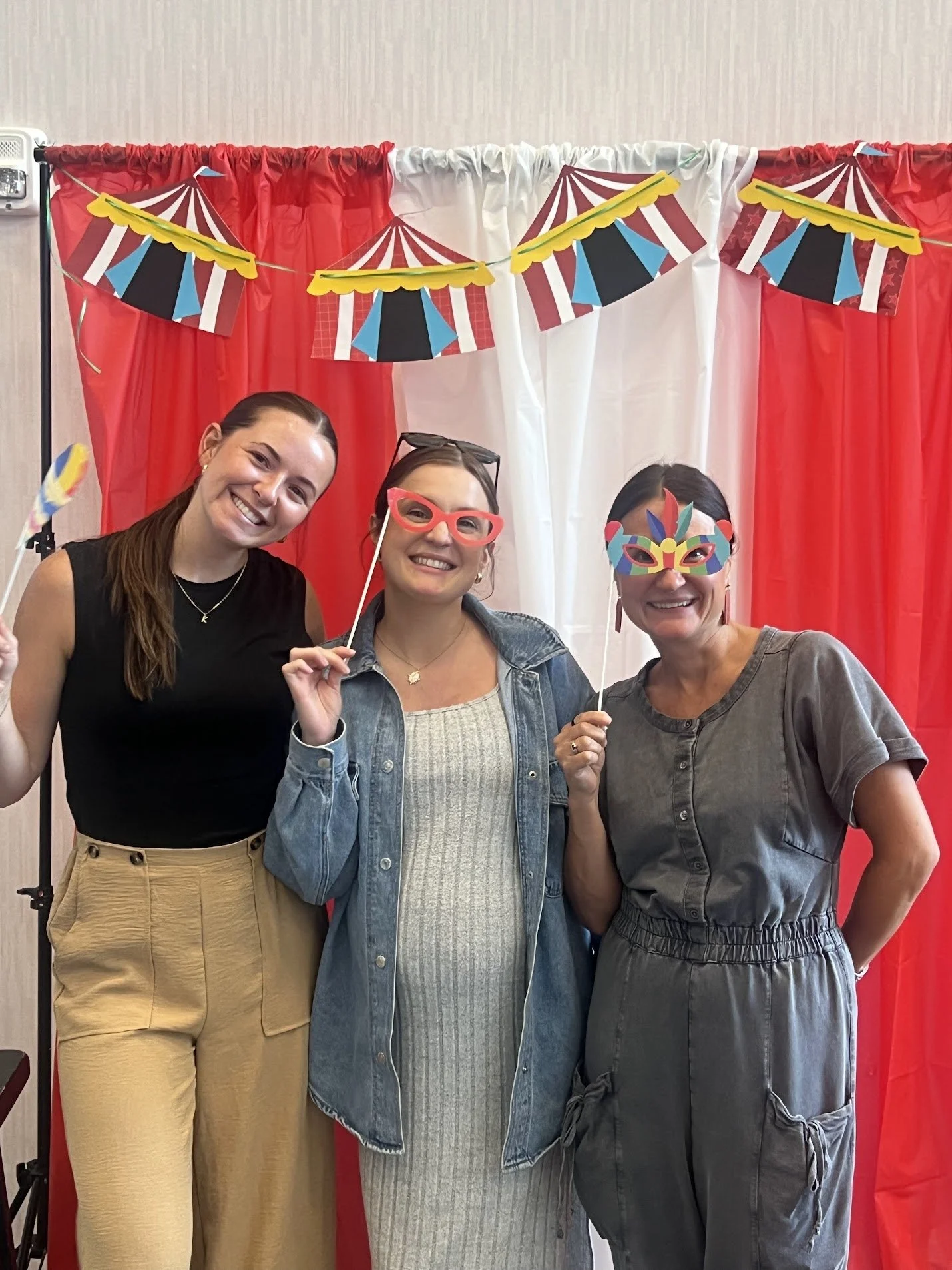 Three women smiling and wearing colorful masks, standing in front of a carnival-themed red and white backdrop with decorative banners.