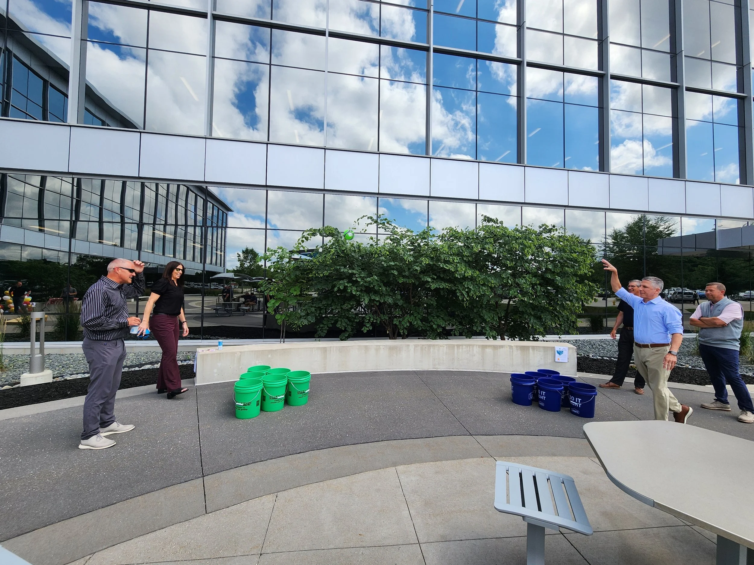 A group of people playing cornhole outdoors in front of a modern glass building.
