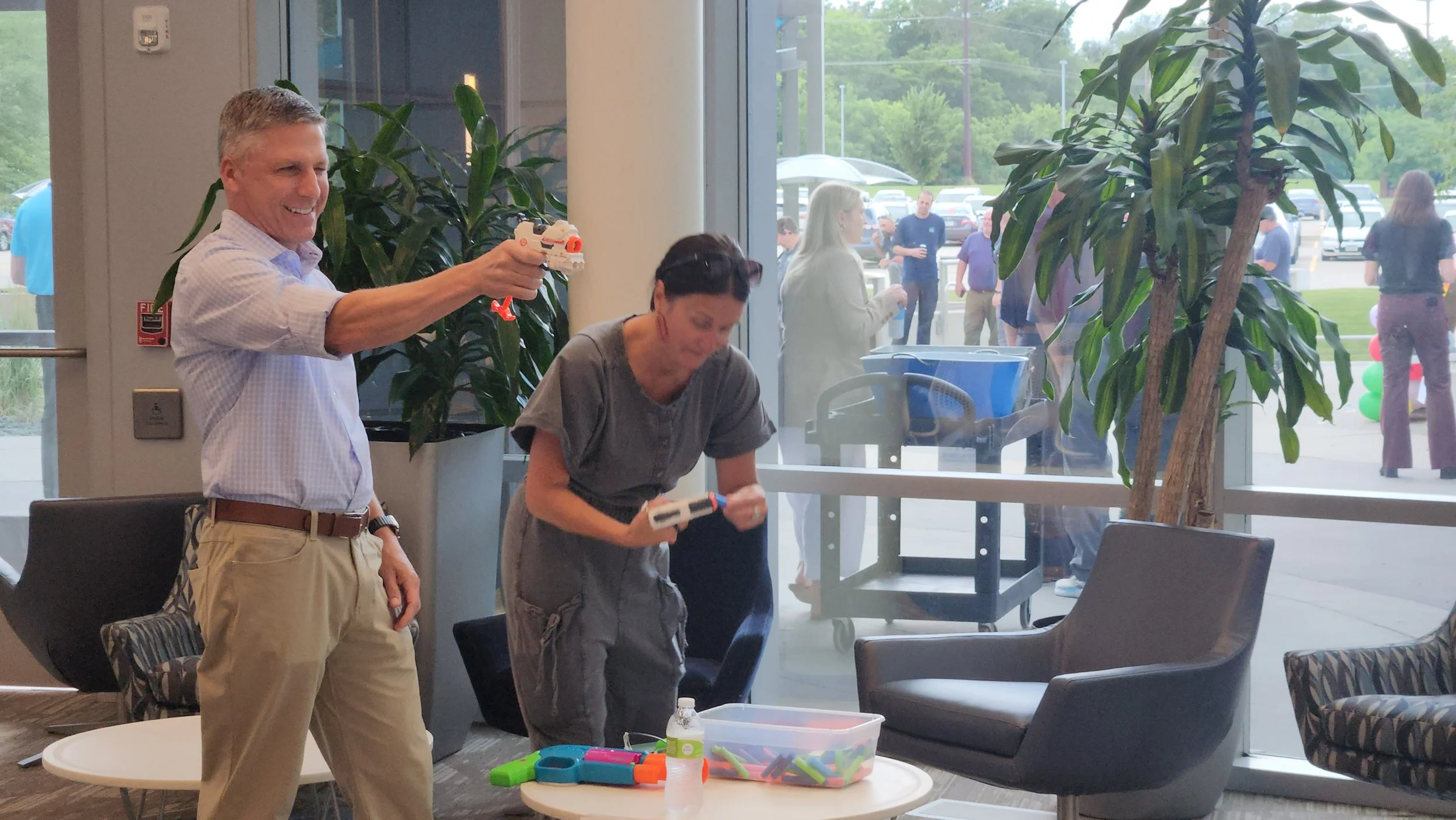 Two adults are inside a building, playing with toy guns and toy foam darts, smiling and laughing. Outside the window, a group of people are gathered in a parking lot.