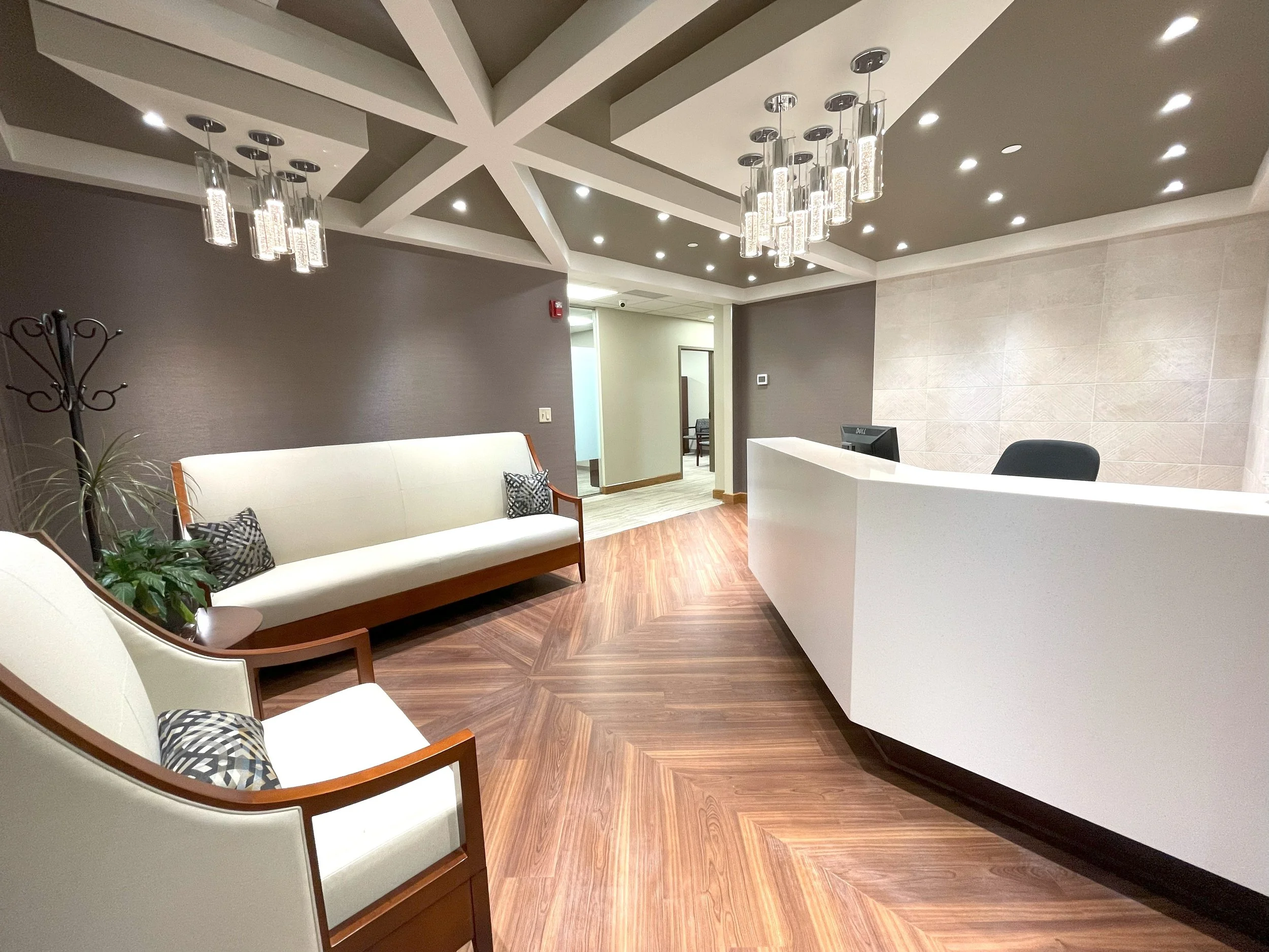 Empty modern reception area with white curved desk, black chair, beige sofa with patterned pillows, armchair, and decorative lighting fixtures overhead.