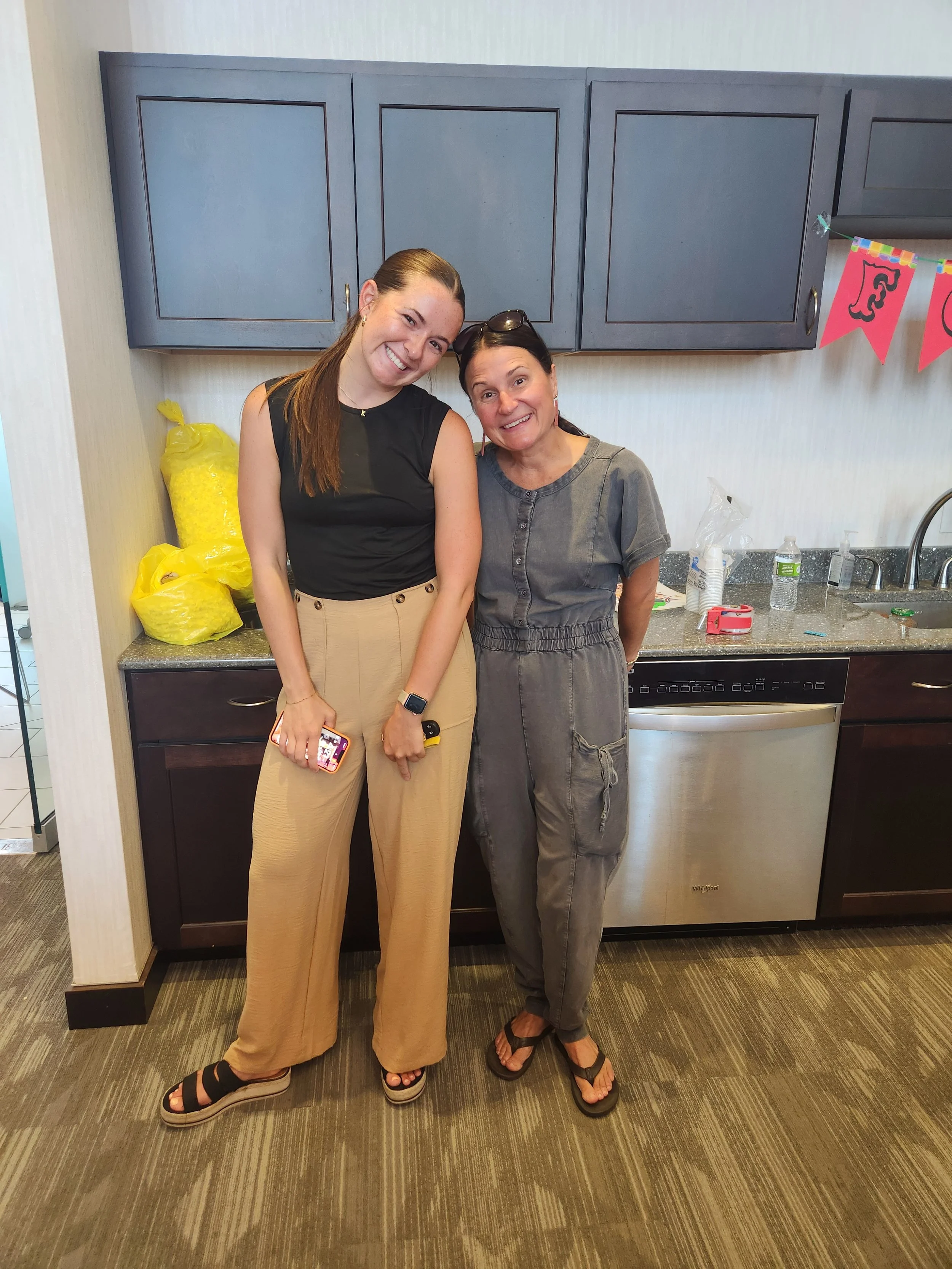 Two women stand close together in a kitchen, smiling at the camera. One woman is holding a phone, and there are yellow bags on the counter behind them, along with a water bottle and some decorations on the wall.