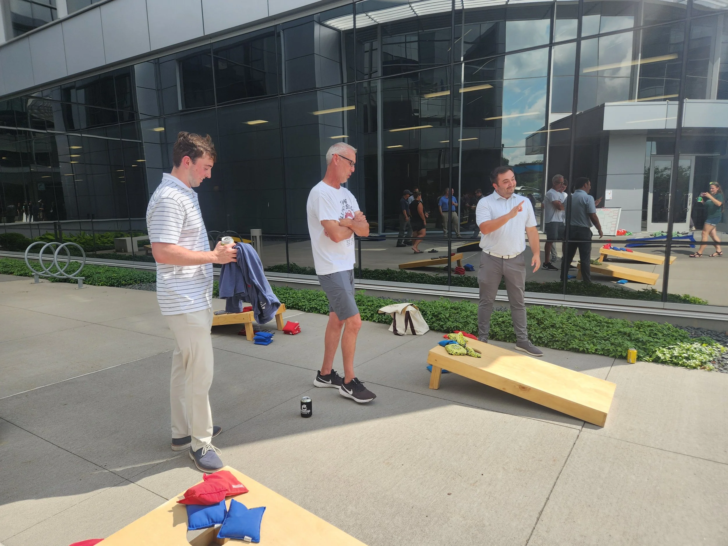 Three men playing cornhole outdoors near a modern glass building. One man is holding a drink, another is preparing to throw, and the third is explaining or instructing. Cornhole boards and bags are visible.