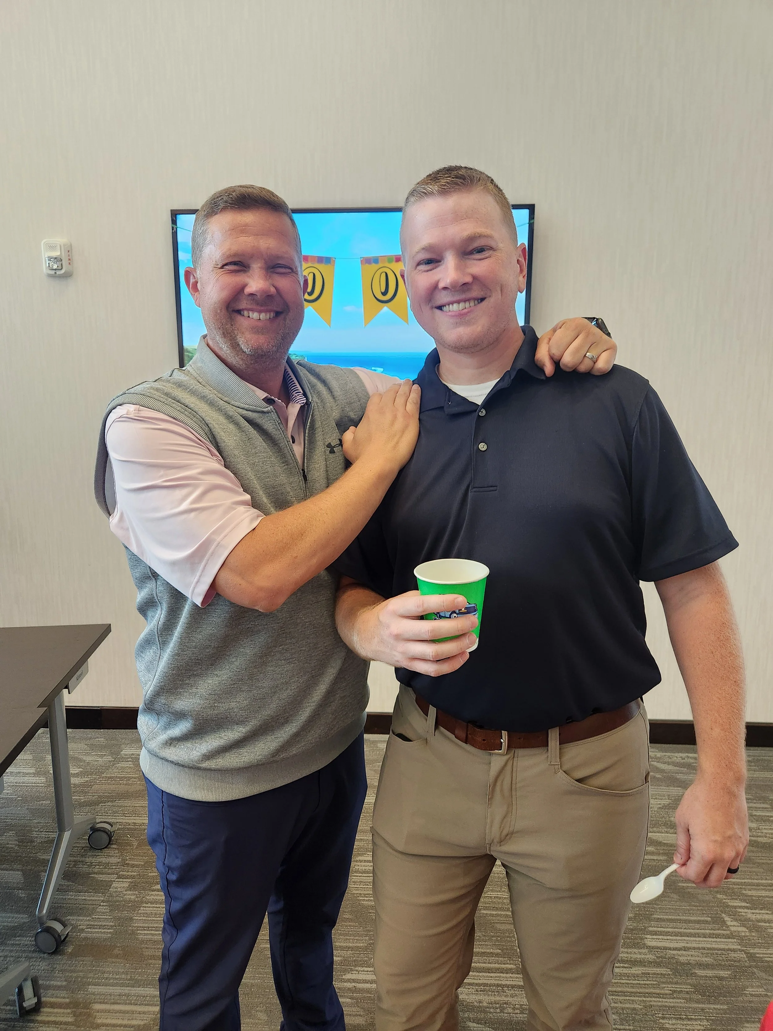 Two smiling men with arm around each other, holding a green cup and a white spoon, standing in a room with a TV behind them displaying a yellow banner. One man is wearing a pink shirt with a gray vest, and the other is in a black polo shirt and khaki