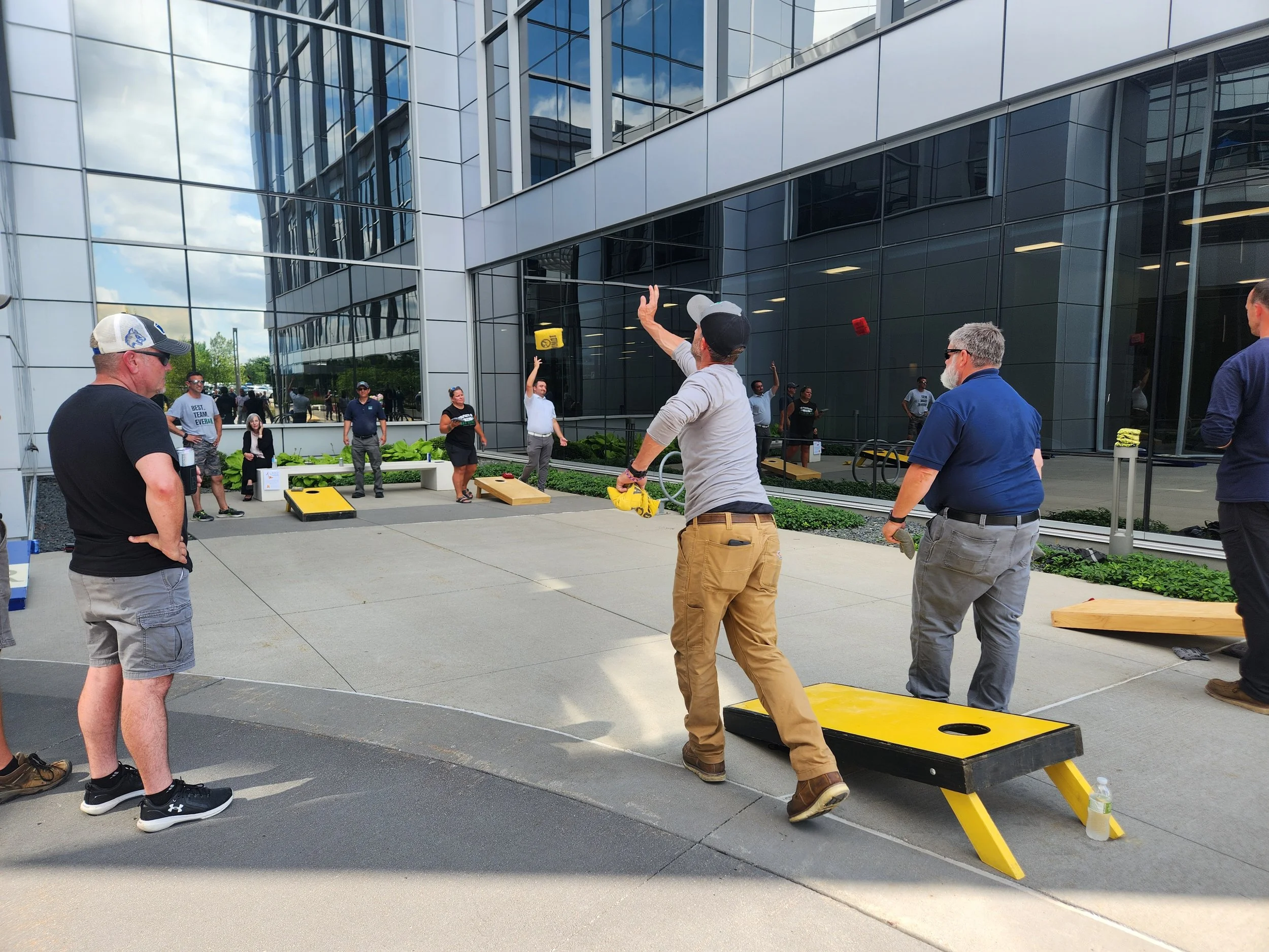 Group of people playing cornhole outside near a modern glass building on a sunny day.