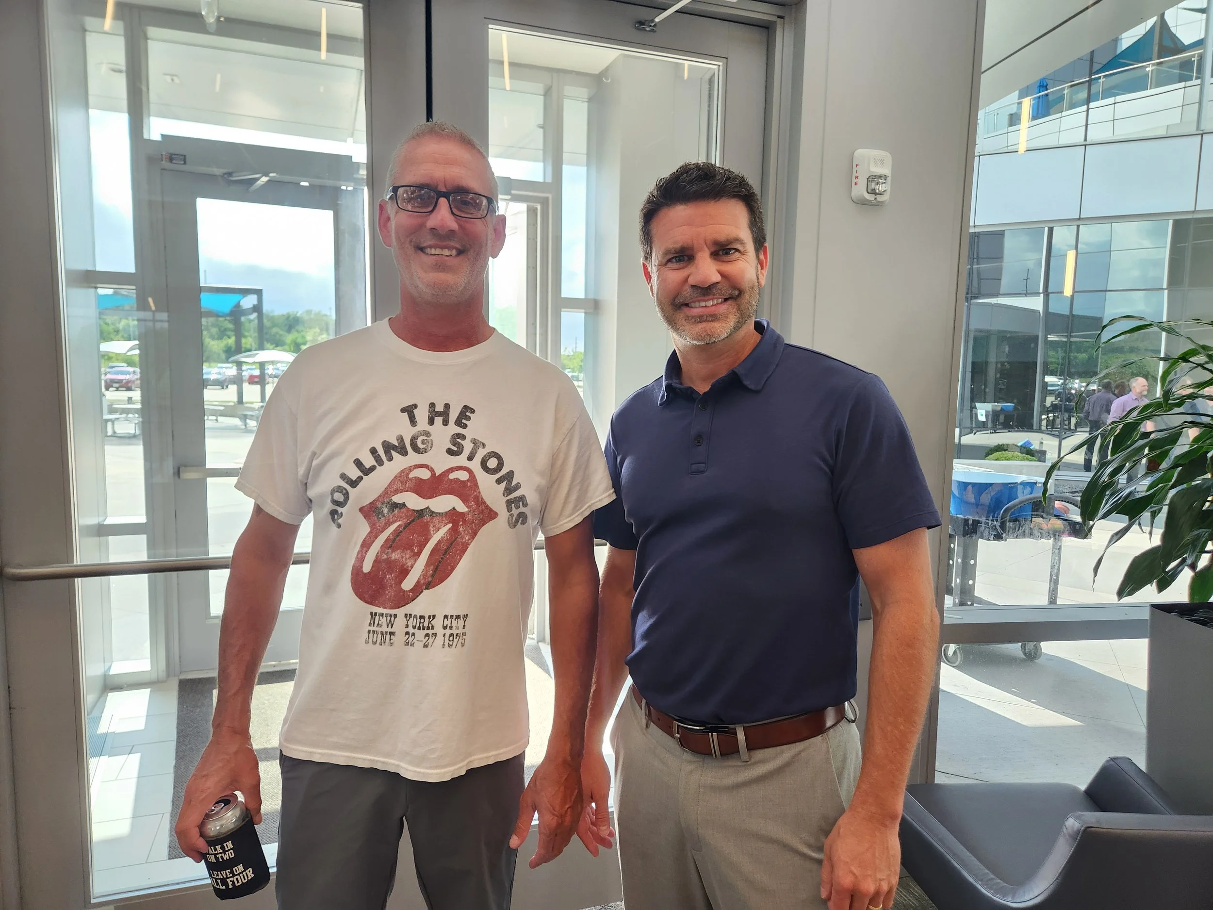 Two men standing inside a building near glass doors, smiling at the camera. One is wearing a Rolling Stones t-shirt, and the other is in a navy polo shirt. Outside, cars and a few people are visible.