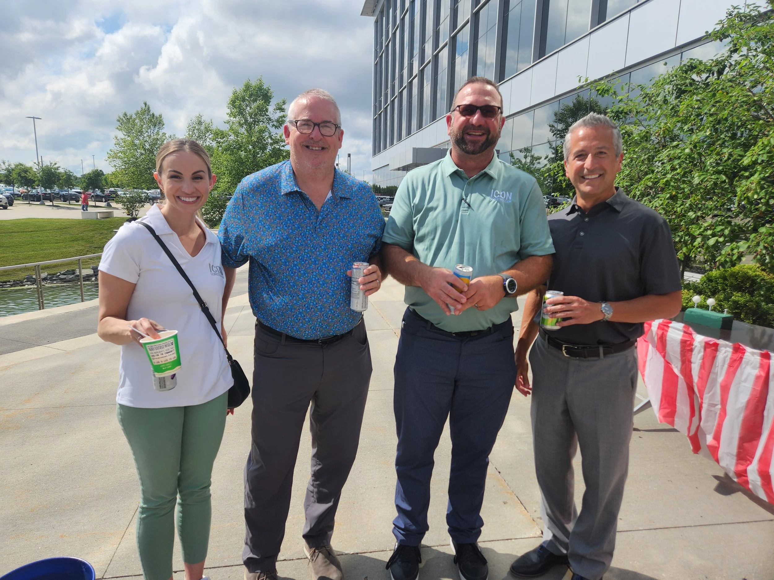 Four people standing together outdoors, smiling and holding drinks, in front of a modern building with trees and a parking lot in the background.