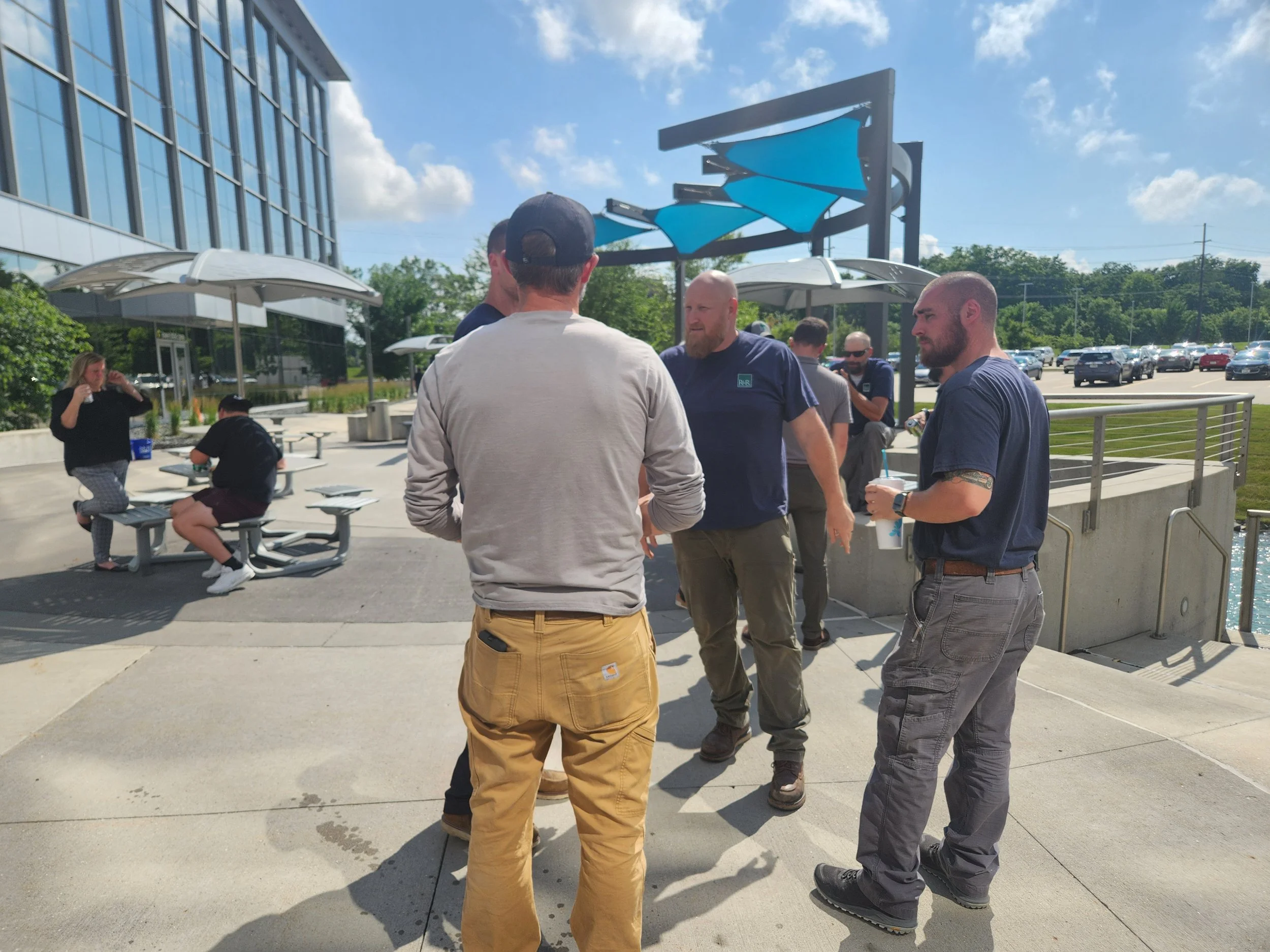 Group of five men standing and talking outside near a modern building with glass windows and a parking lot, with some women sitting on benches in the background.