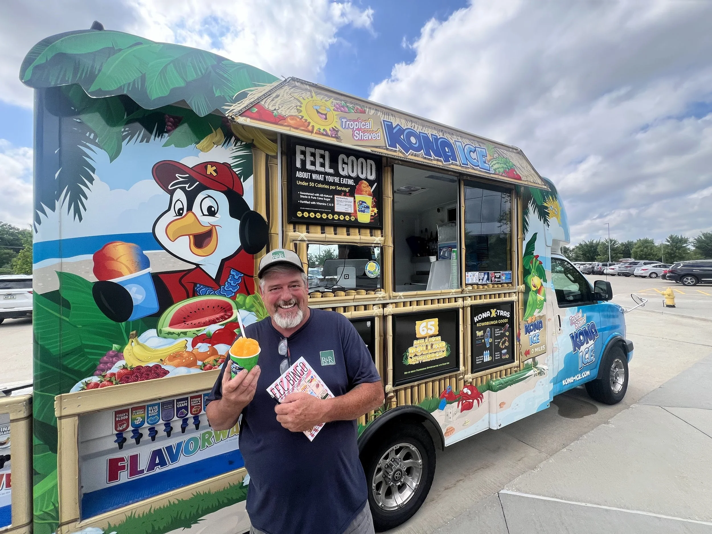 A man smiling and holding a rainbow-colored snow cone with a food truck decorated with tropical-themed graphics in the background.