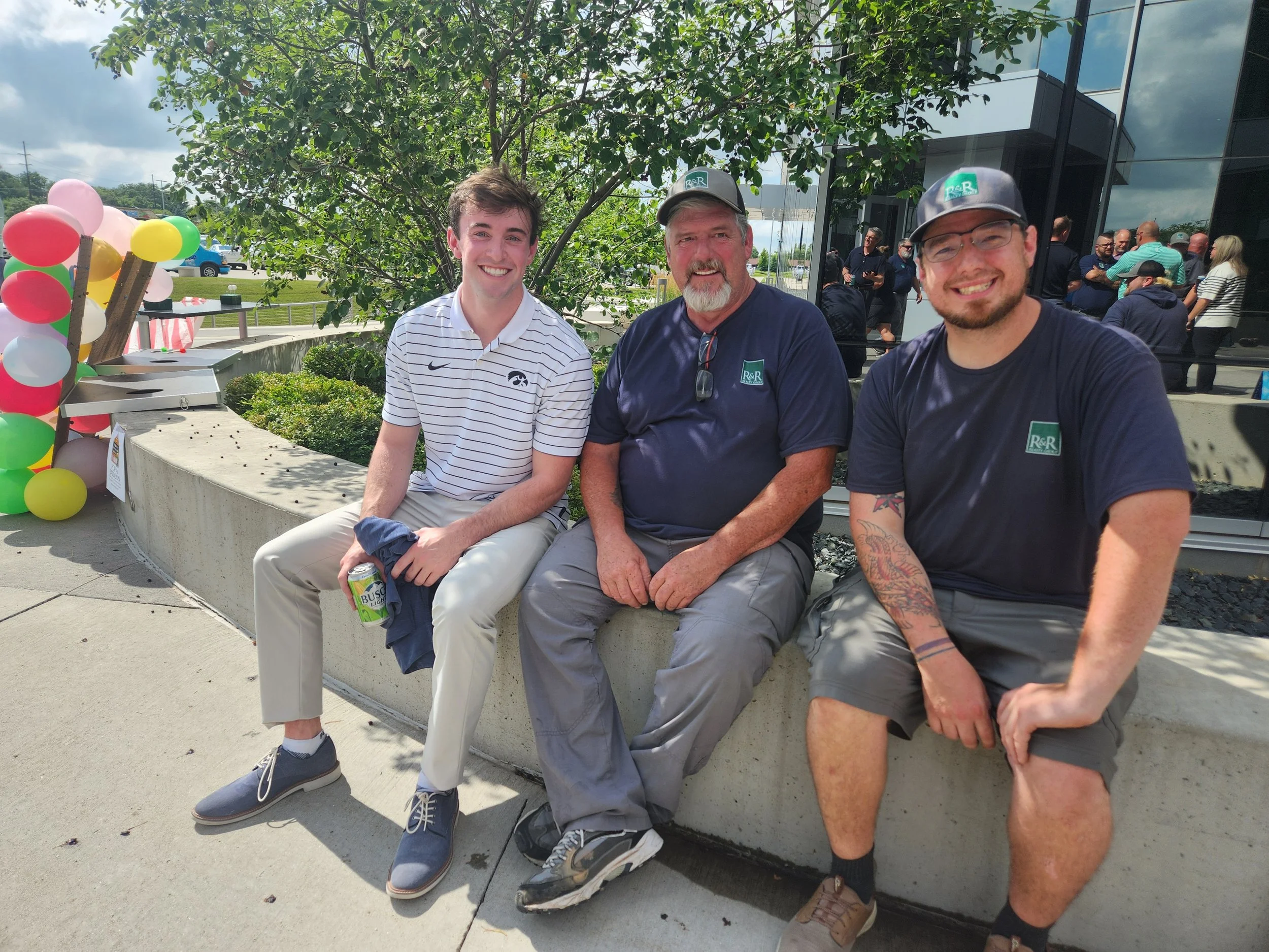 Three men sitting on a concrete ledge outdoors, smiling at the camera during a celebration event with balloons and a crowd in the background.