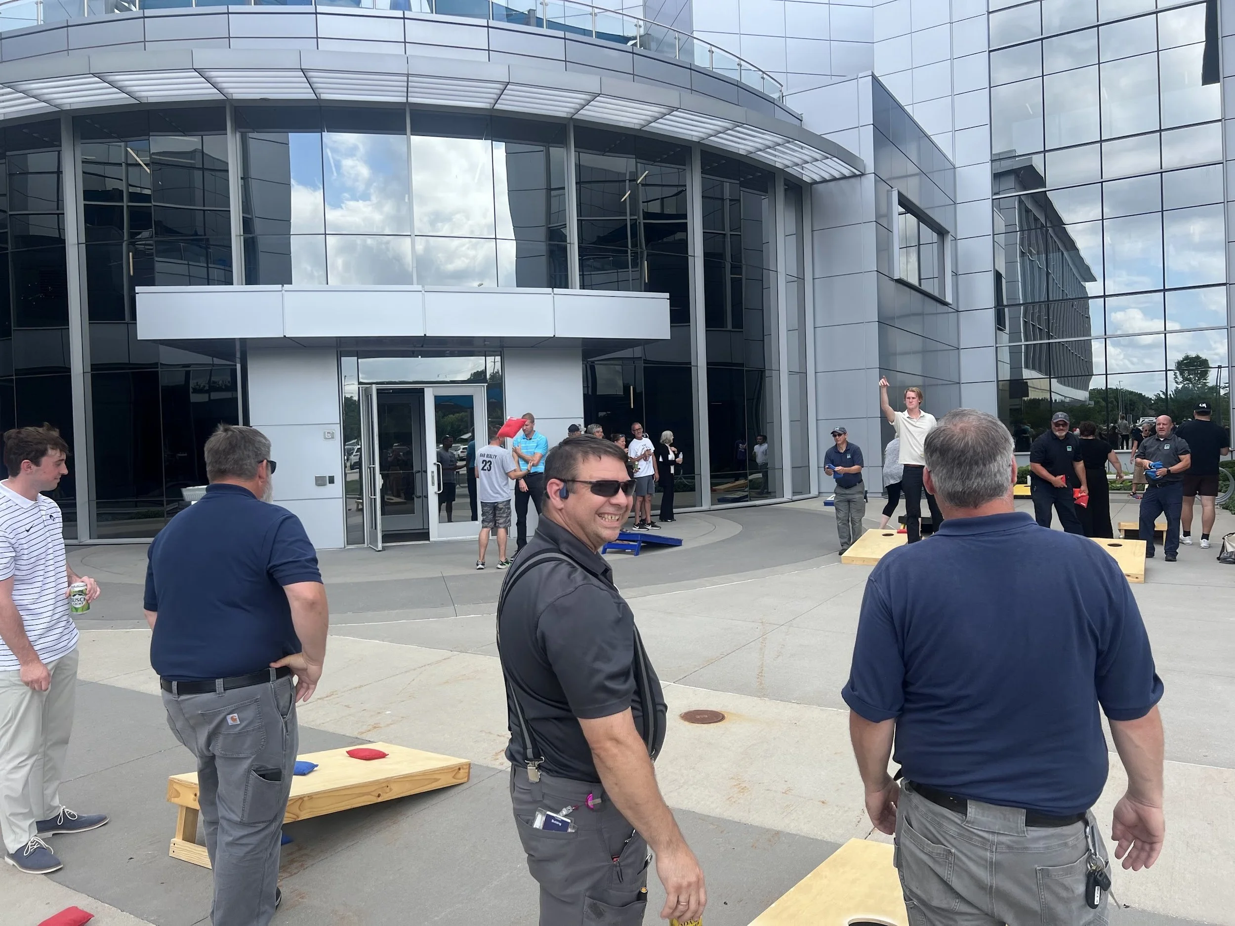 People gathered outside a modern glass building participating in a cornhole game, with some playing and others watching. A man in a white shirt is standing on a wooden platform, preparing to throw a bean bag.