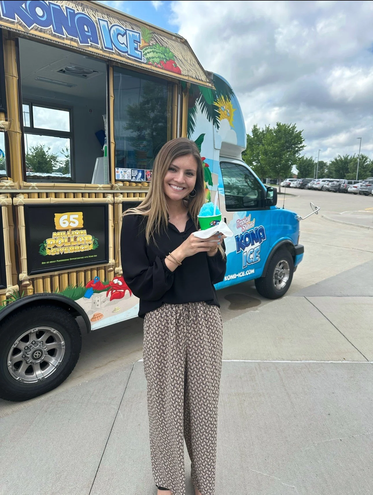 Woman smiling and holding a blue Hawaiian shaved ice dessert in front of a Kona Ice truck decorated with tropical graphics.