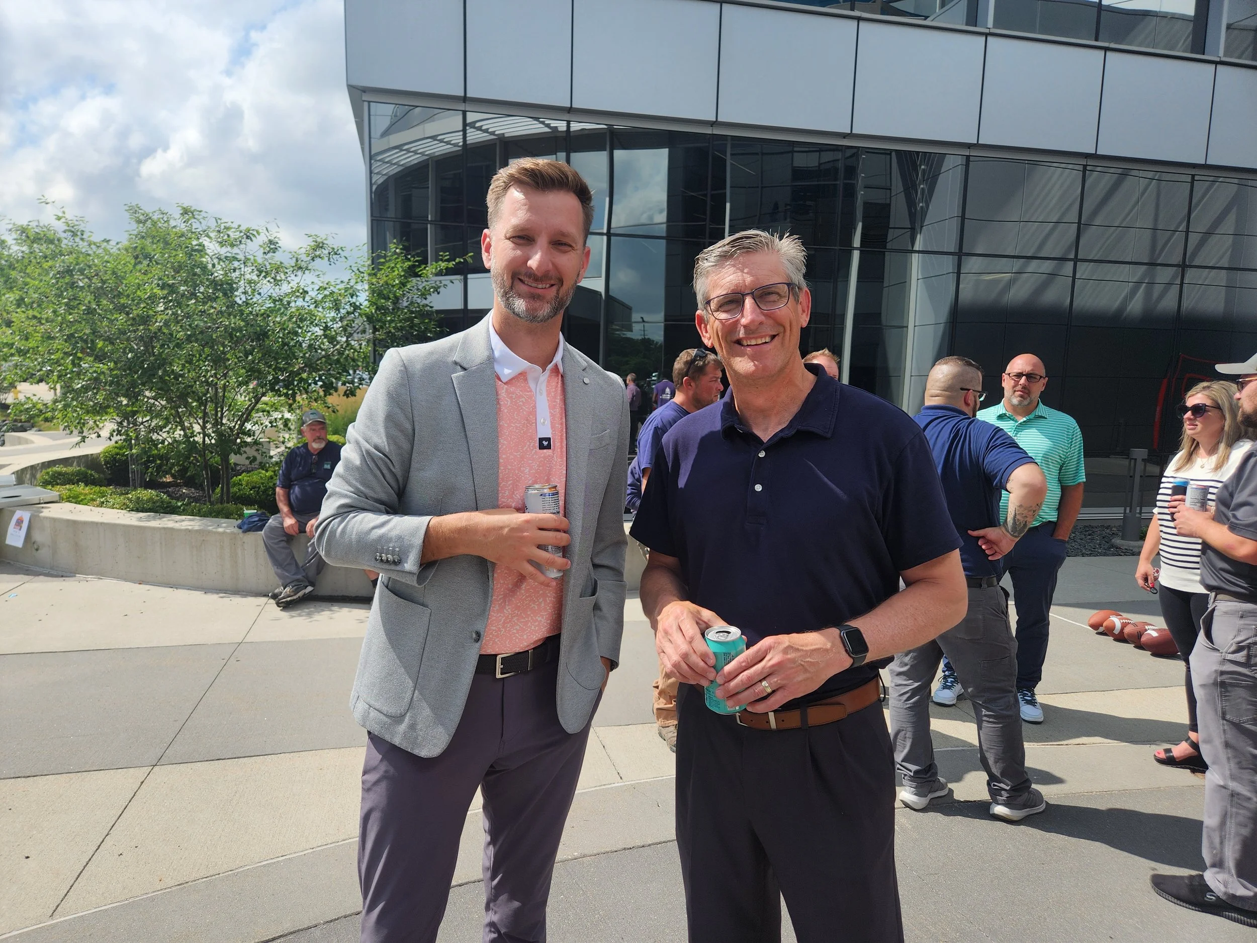 Two men are smiling and holding canned drinks while standing outdoors in front of a modern glass building with a group of people in the background, some sitting and some conversing.