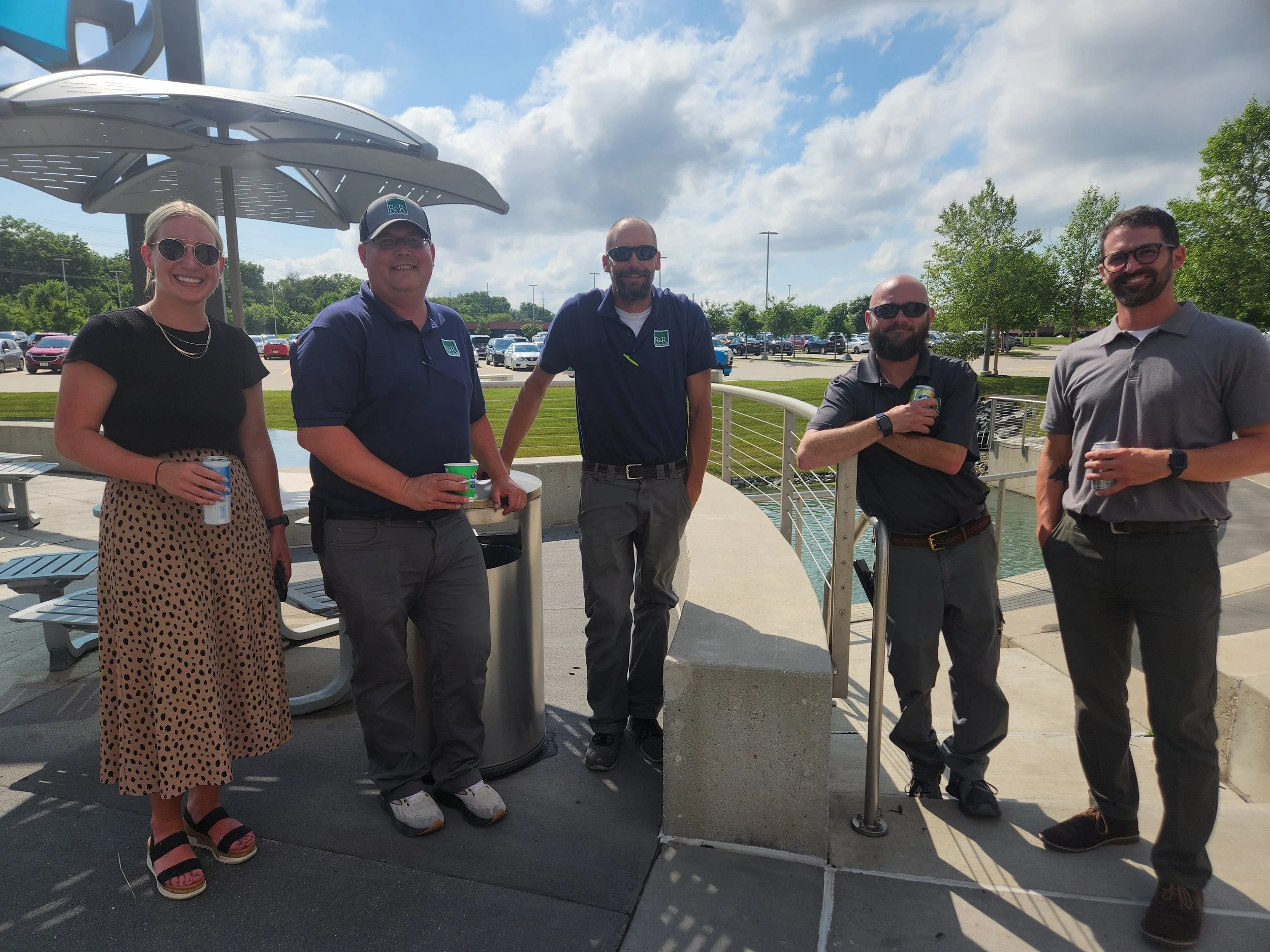 Group of six people standing outdoors near water and parking lot, smiling and holding drinks, on a sunny day with partly cloudy sky.