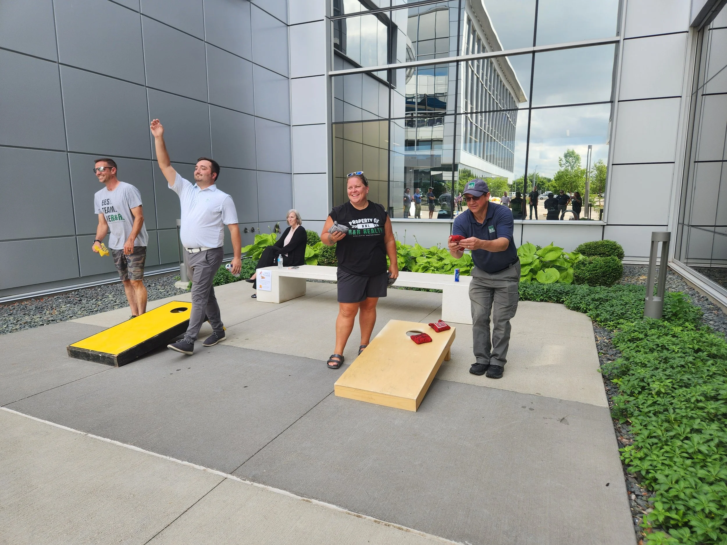 People playing cornhole outside a modern building with large glass windows; one person is mid-throw, others are watching or preparing their turn.