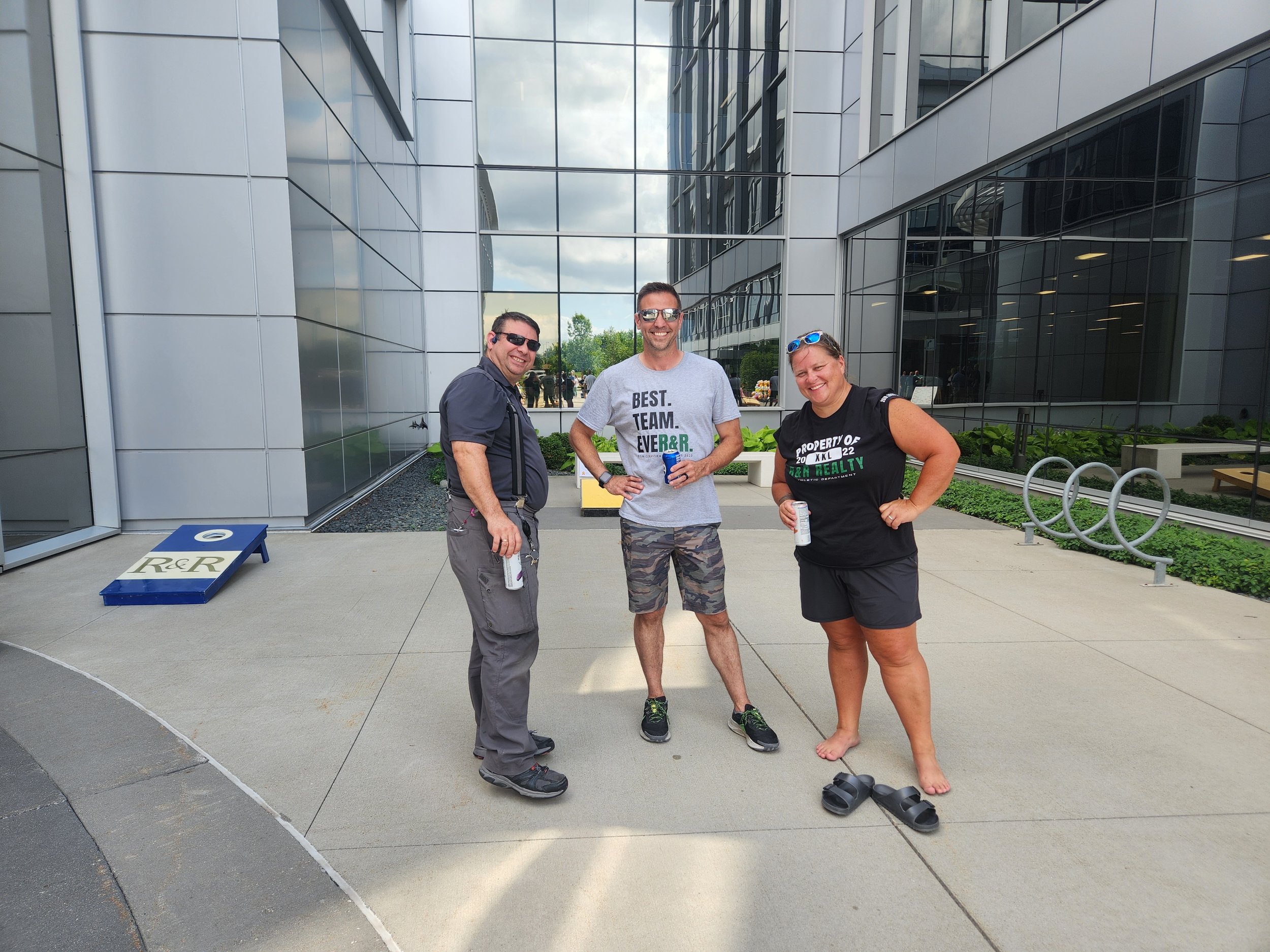 Three people standing outside a modern glass building, smiling, holding drinks, with sandals on the ground, and bicycle racks in the background.
