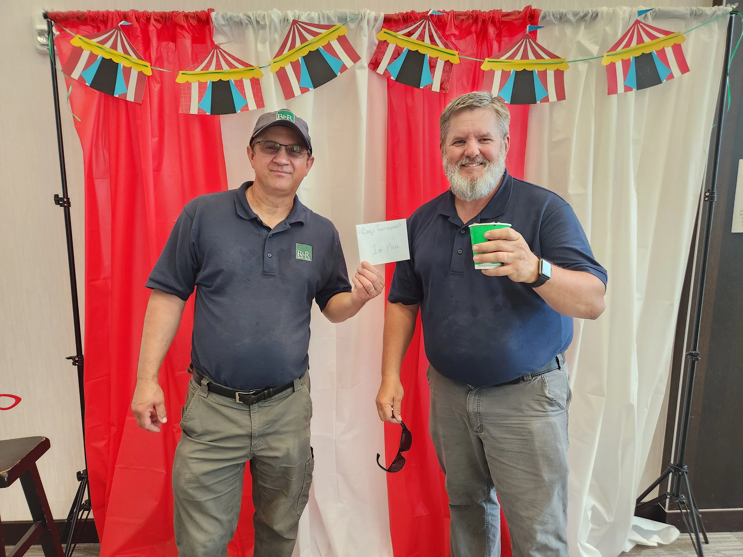 Two men standing together in front of a red and white curtain backdrop with circus tent decorations, one holding a note that says 'Bag Tournament 1st Place', and the other holding a green cup. Both men are wearing navy blue polo shirts and gray pants