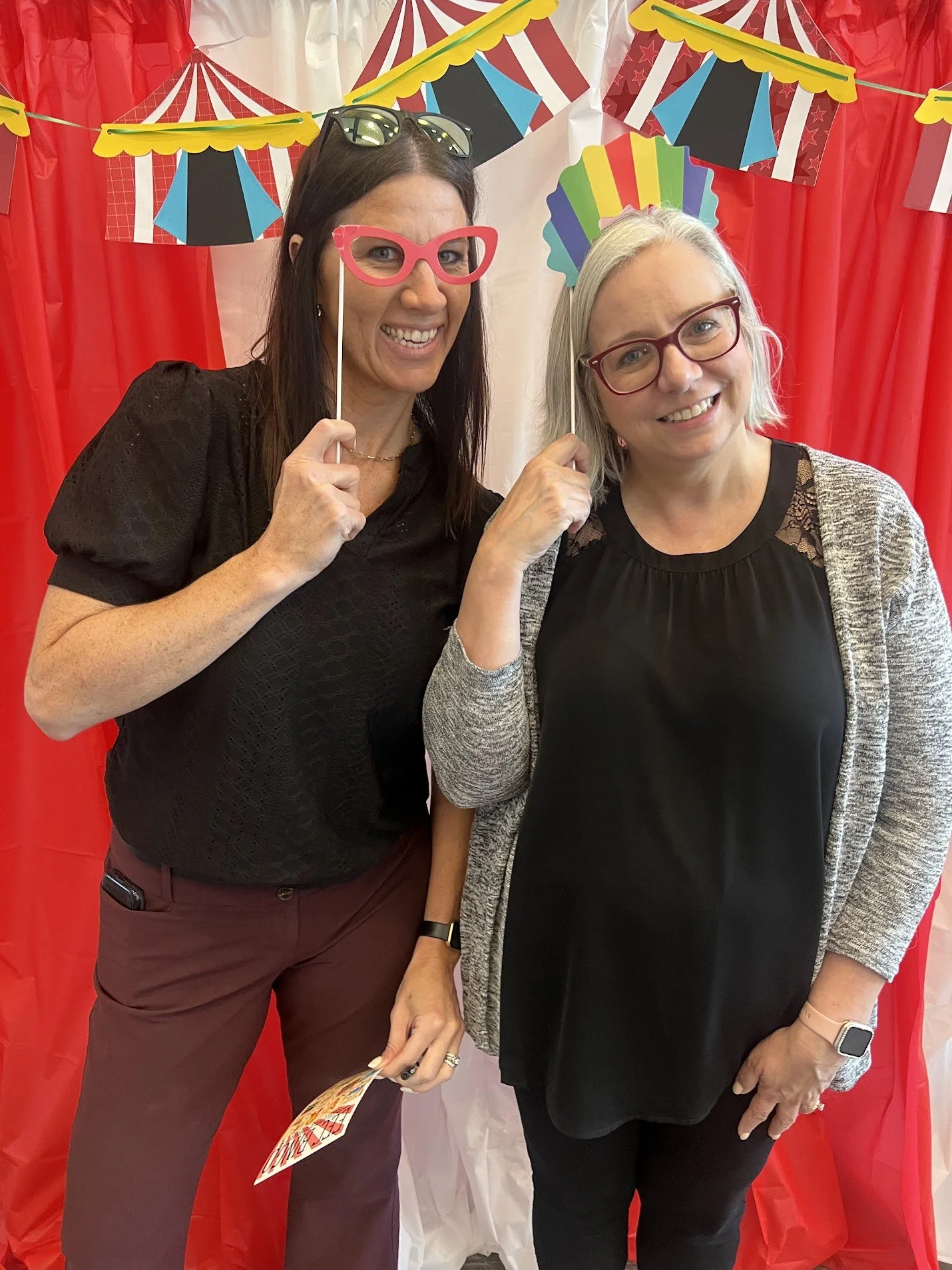 Two women at a festive event holding colorful costume props, with red and white curtains and circus-themed banners in the background.