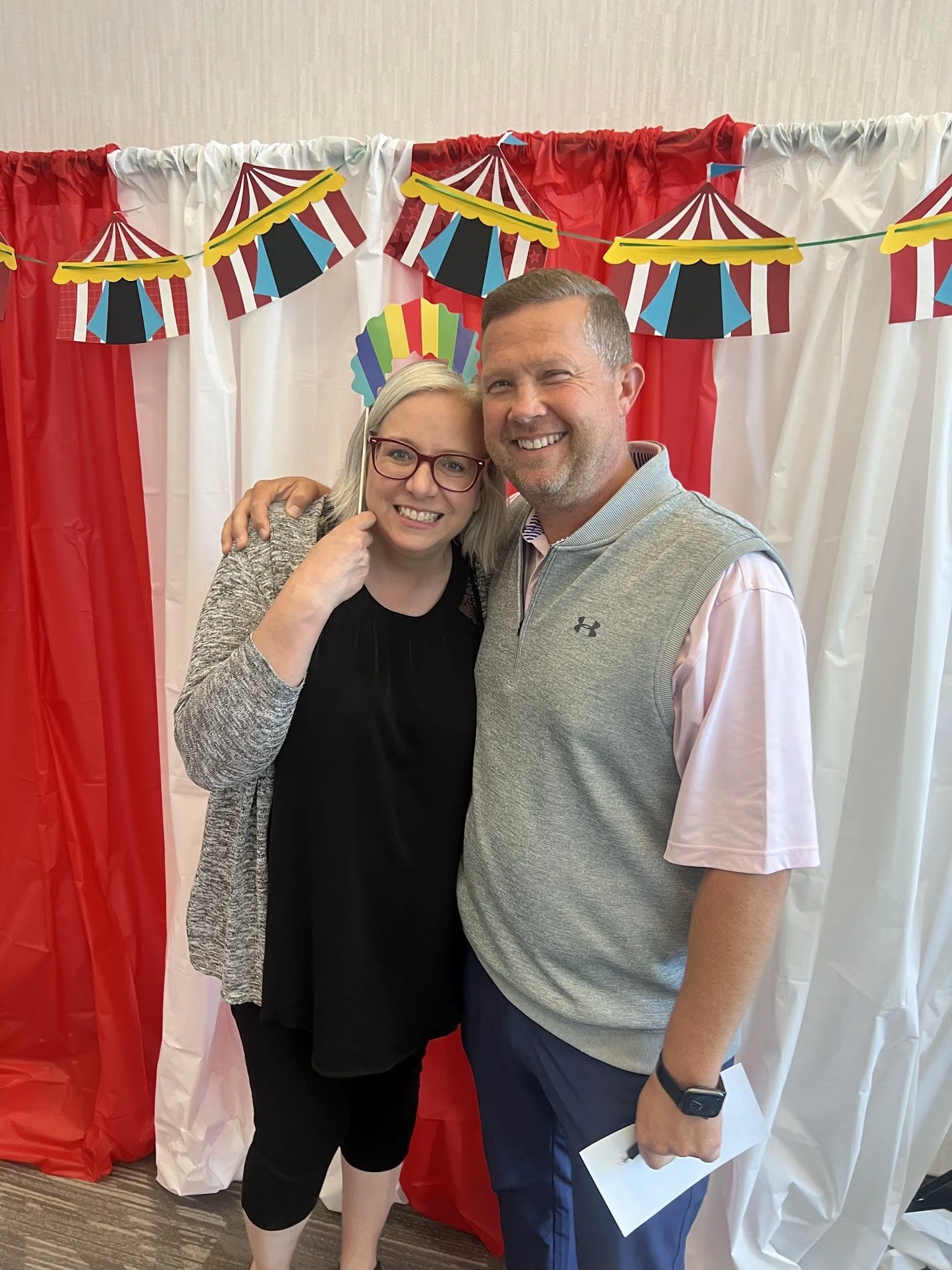 A smiling woman and man standing close together in front of a colorful circus-themed backdrop with red, white, and multicolored decorations.
