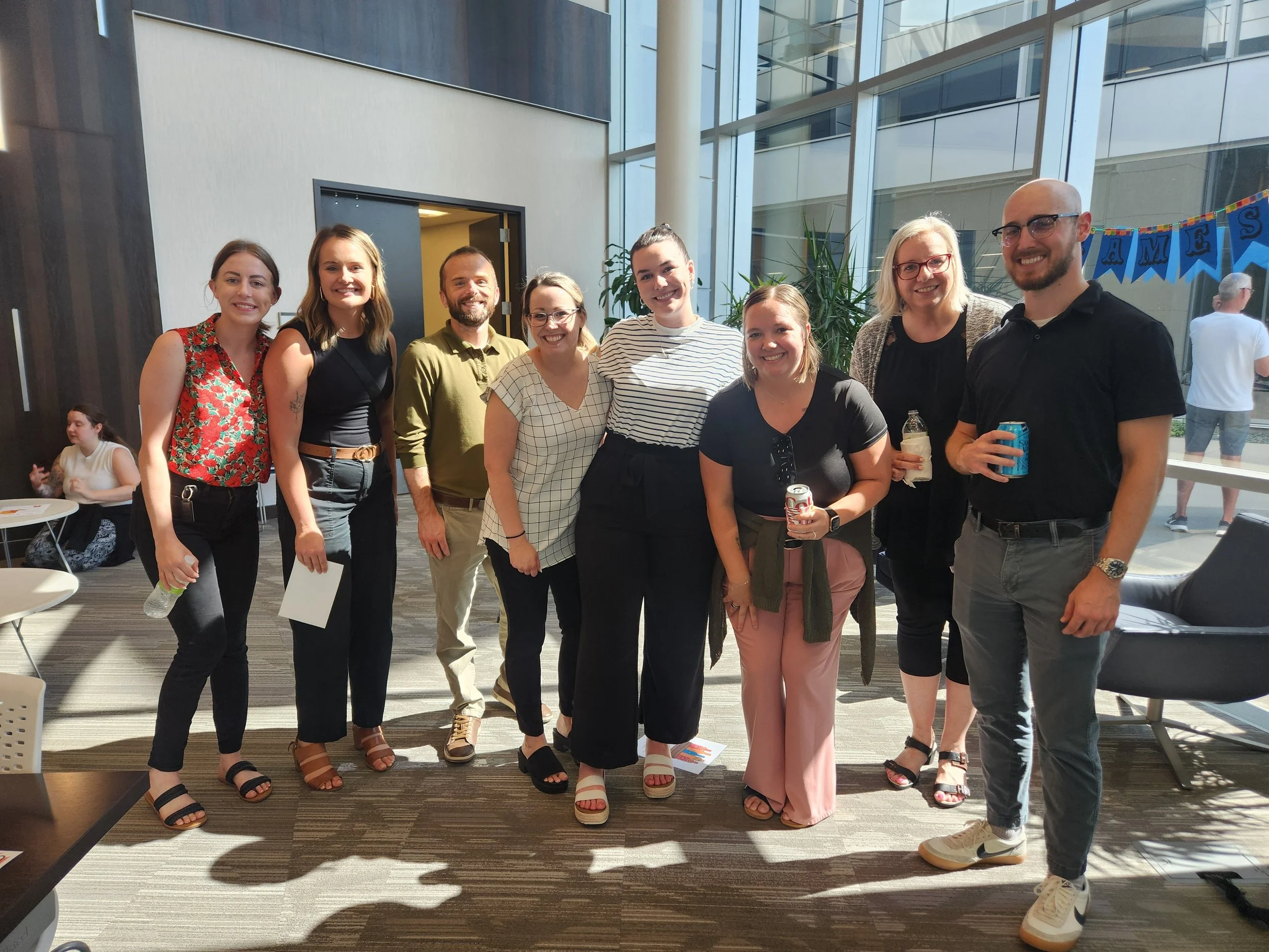 Group of nine people standing inside a modern building, smiling for the photo, with sunlight streaming in from large windows behind them.