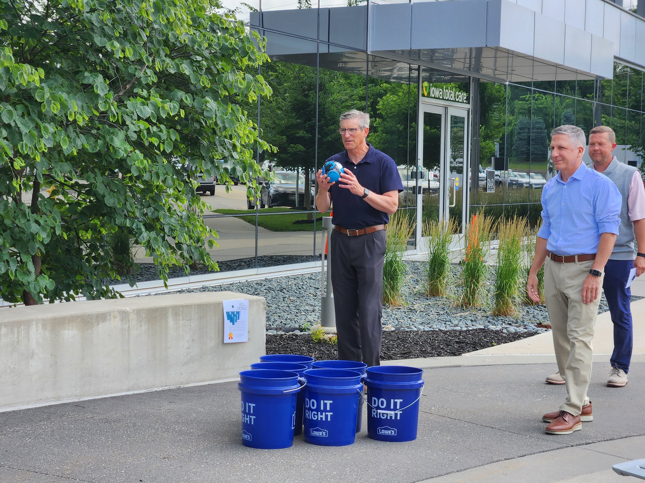 Three men standing outside near a modern building with reflective glass walls, one man holding a blue ball, next to blue buckets with the words 'DO IT RIGHT' on them, and a sign attached to a concrete barrier that says 'Big Pong' and has a drawing of
