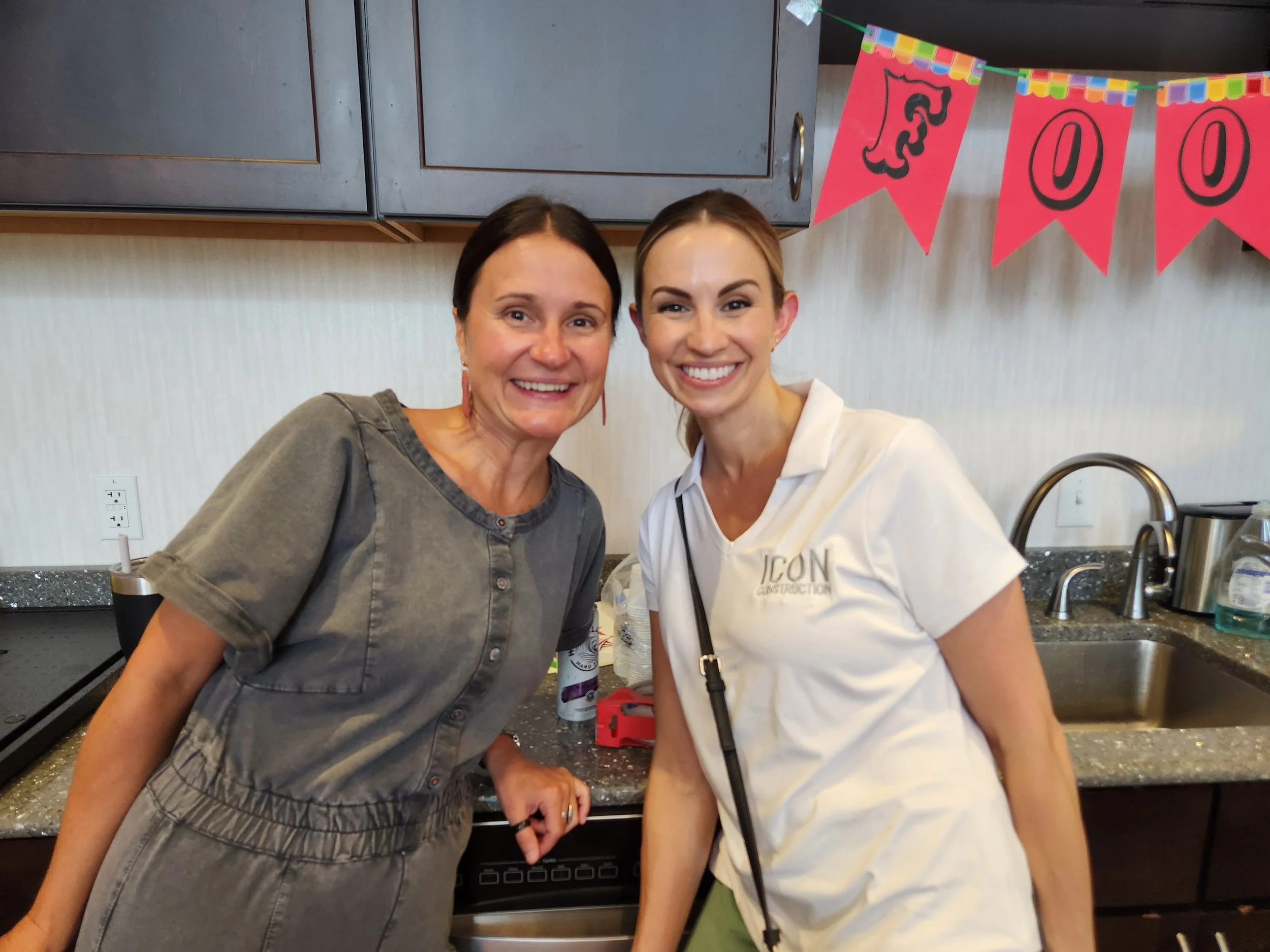 Two women standing in a kitchen, smiling at the camera. One woman is wearing a gray dress, and the other is wearing a white shirt with the text 'ICON CONSTRUCTION' embroidered on it. There is a pink banner with letters and a black lion on it hanging 