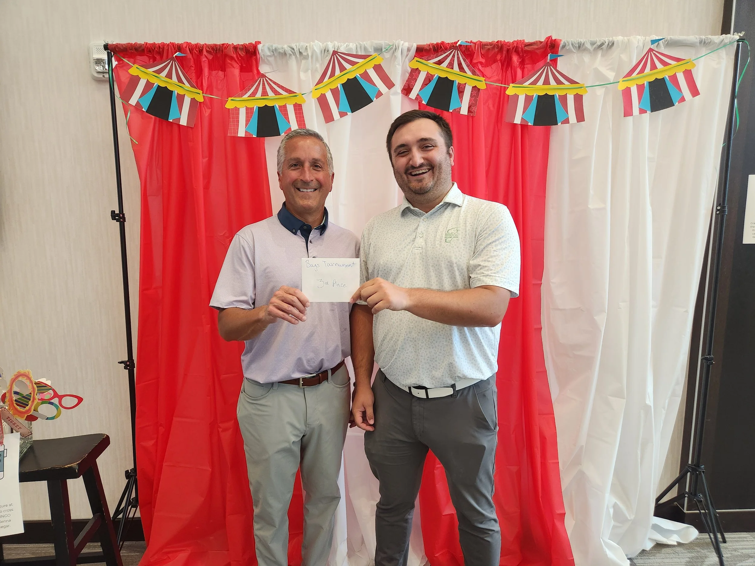 Two men standing in front of red and white curtains with carnival-themed paper banners, smiling. One man holds a note that reads 'Bag Tournament 3rd Place'.