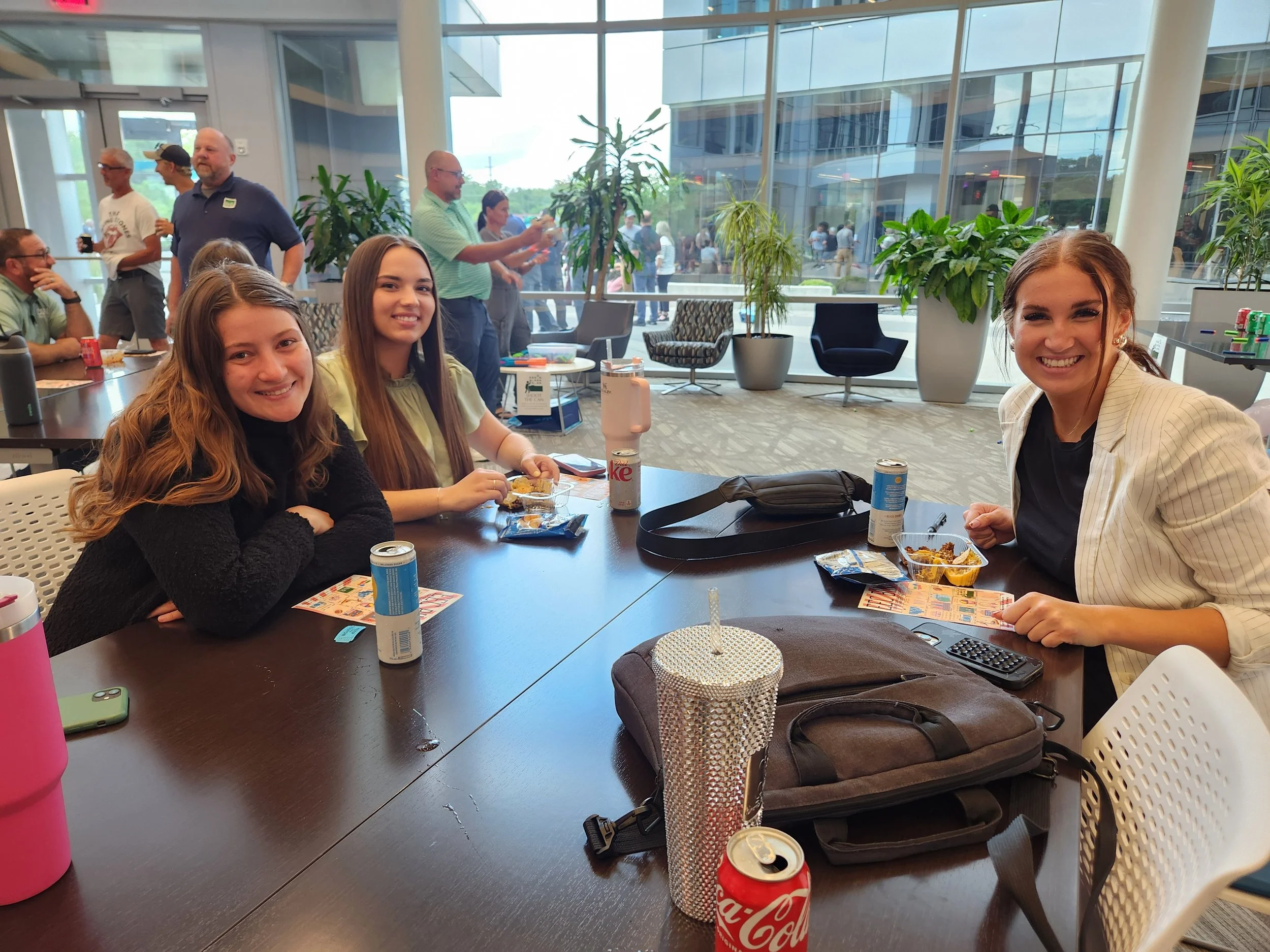 Three young women sitting at a table in a modern indoor space with large windows, enjoying snacks and drinks, while other people are mingling in the background.