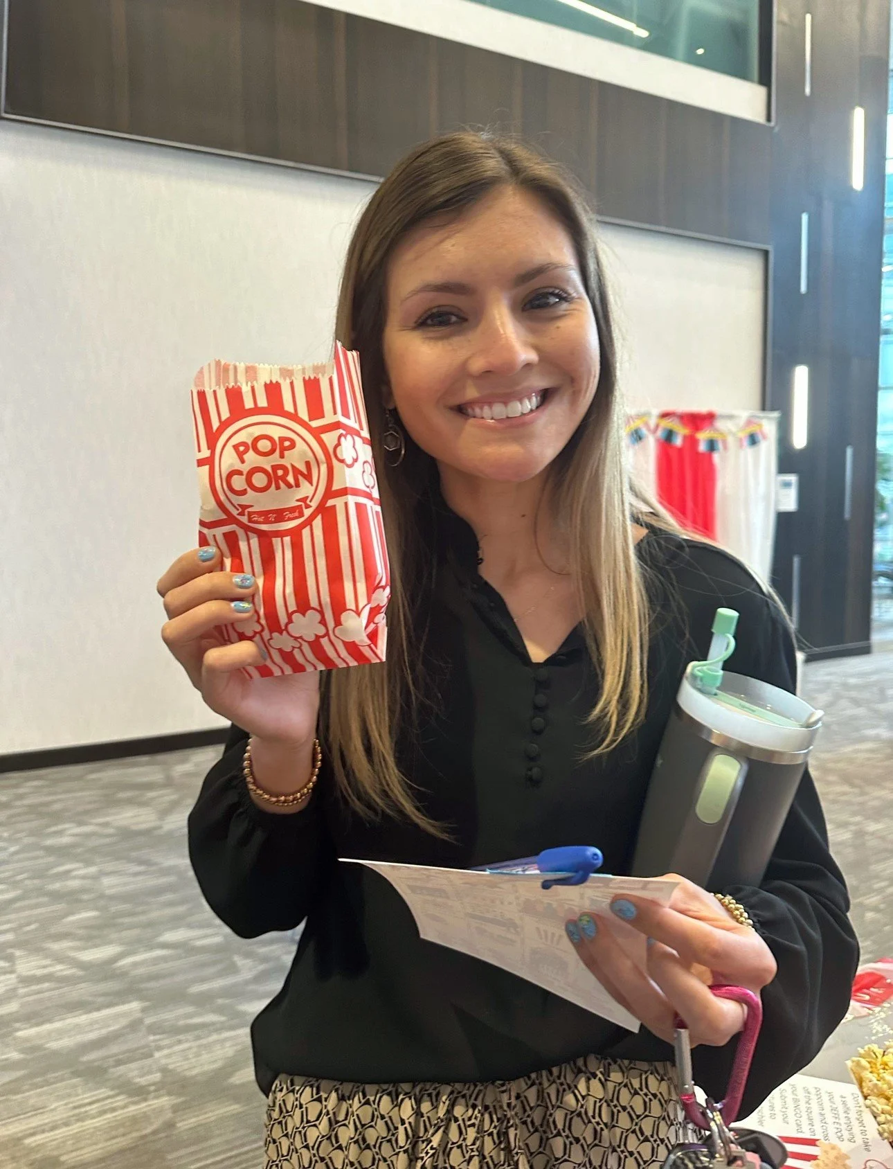 A woman smiling and holding a striped red and white popcorn bag in one hand and a stainless steel tumbler in the other, standing indoors with wood paneling on the wall behind her.
