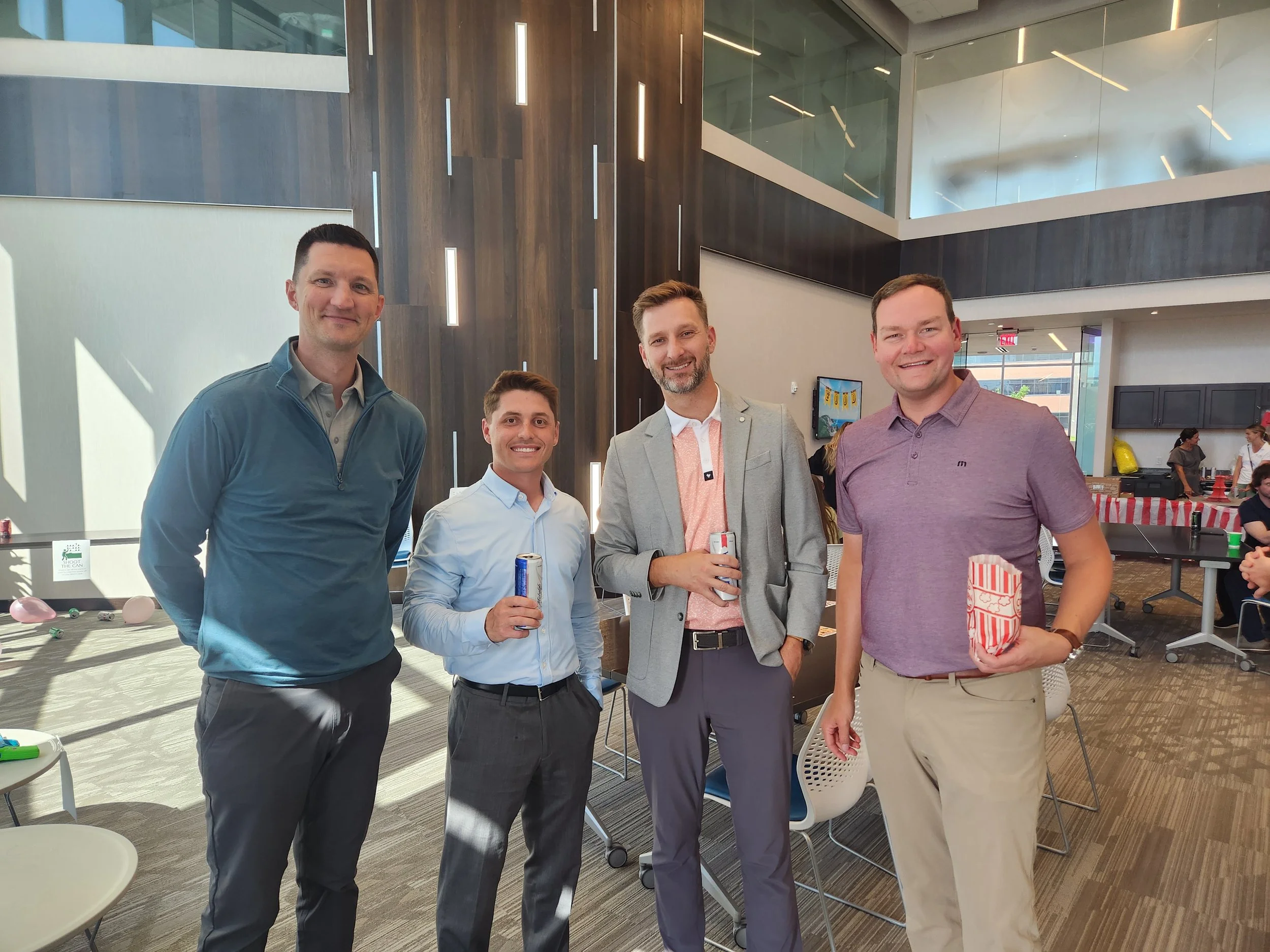 Four men standing together indoors at a social event, holding drinks and smiling at the camera.