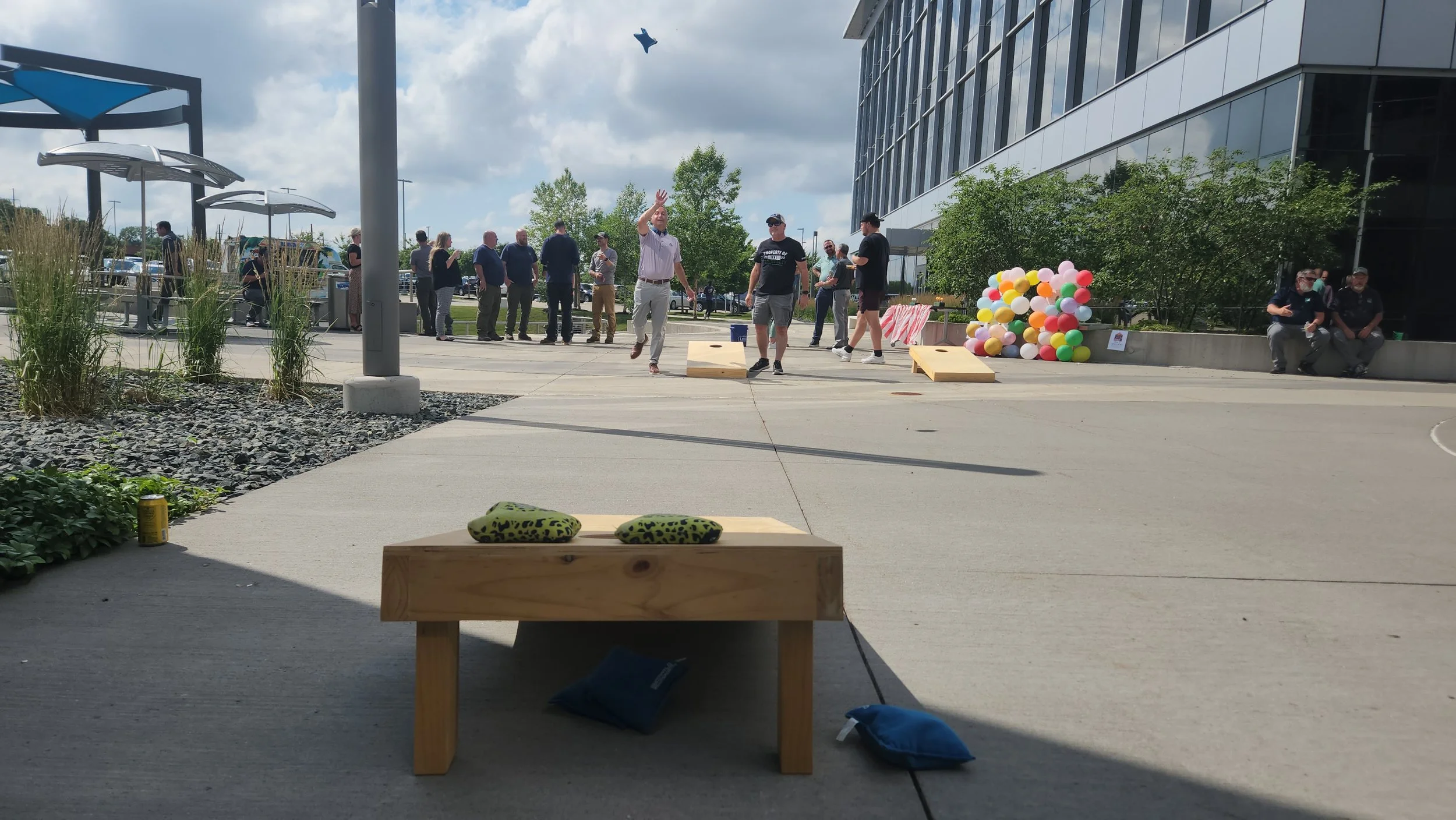 People participating in an outdoor cornhole game near a modern building with balloons and group of people in the background.