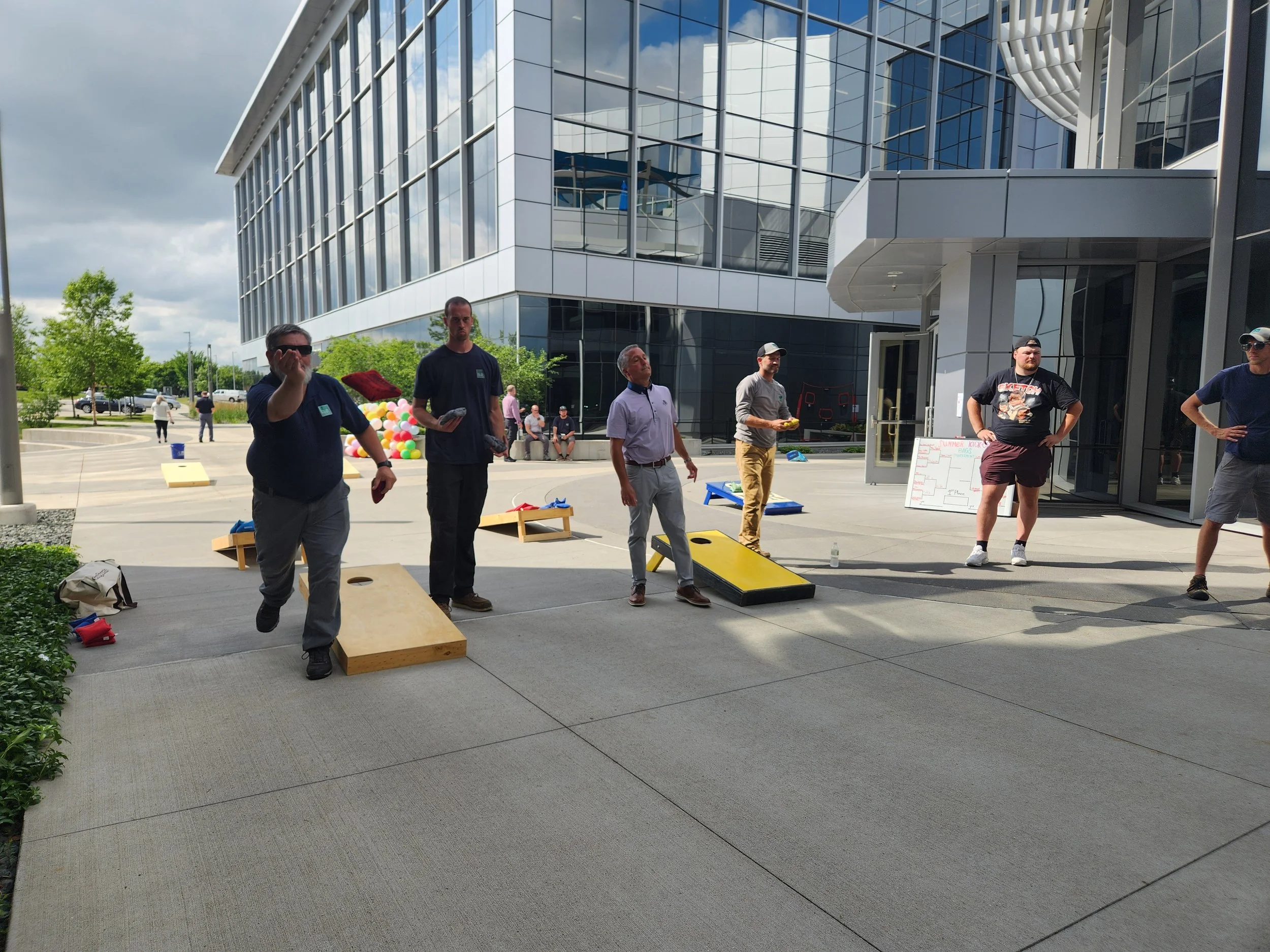 Group of seven men playing cornhole outside in front of a modern glass building during daytime. Some are preparing to toss bean bags into the holes of the wooden boards, with a man in sunglasses and a dark shirt in the foreground, and others standing