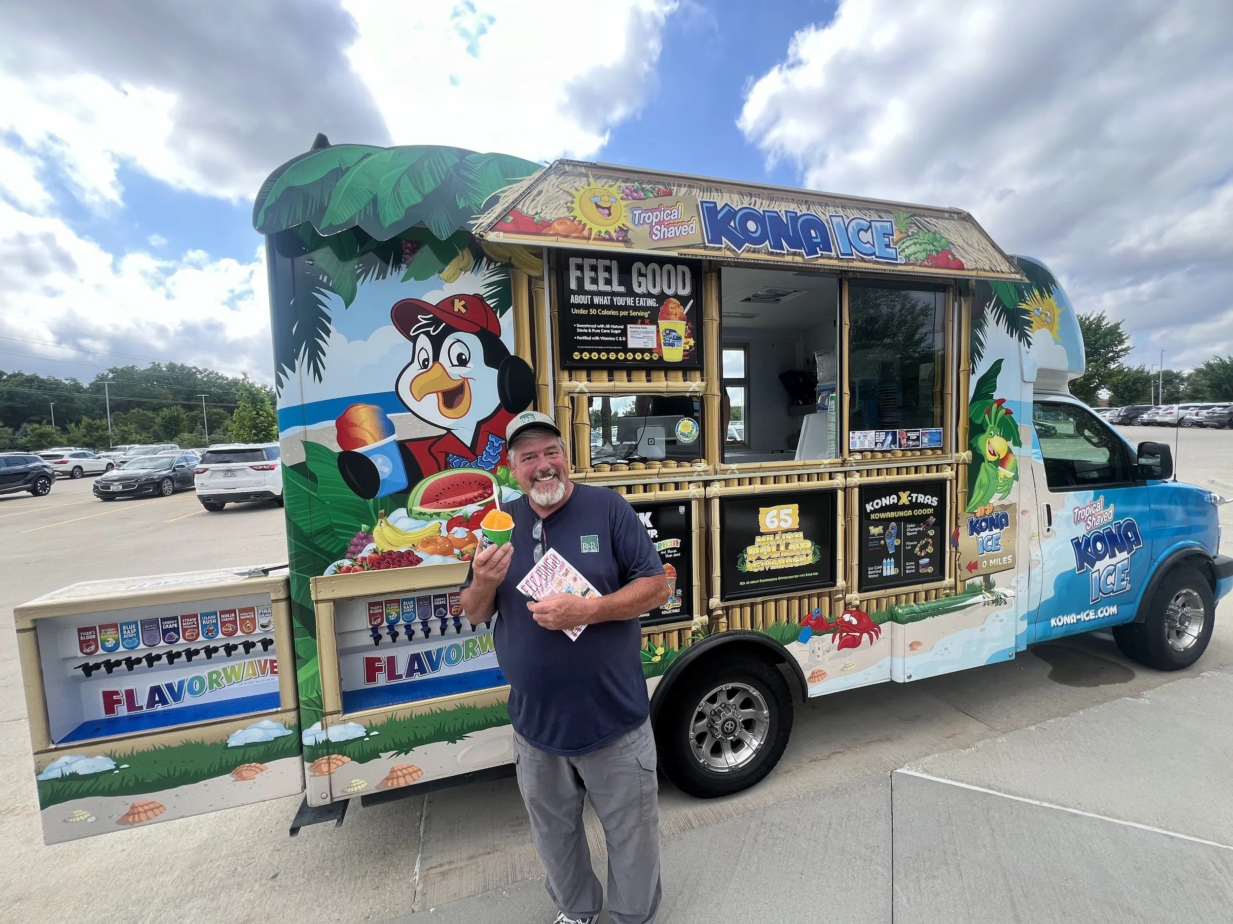 Man smiling and holding a colorful ice cream cone in front of a decorated ice cream truck with cartoon penguin and tropical theme.