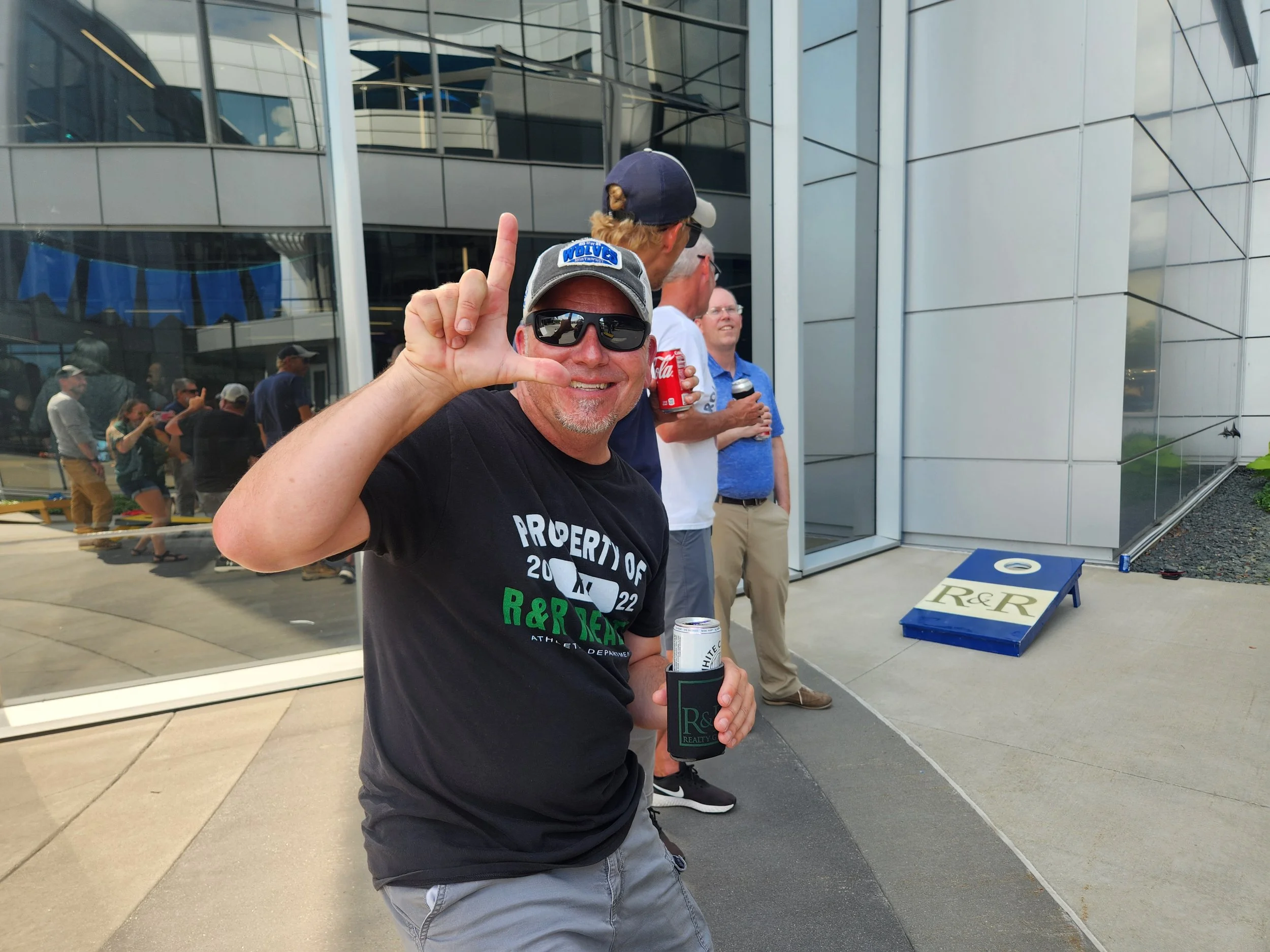 A man smiling and making an 'L' shape with his fingers at an outdoor gathering in front of a modern building. He is wearing sunglasses, a black T-shirt, and holding a drink.