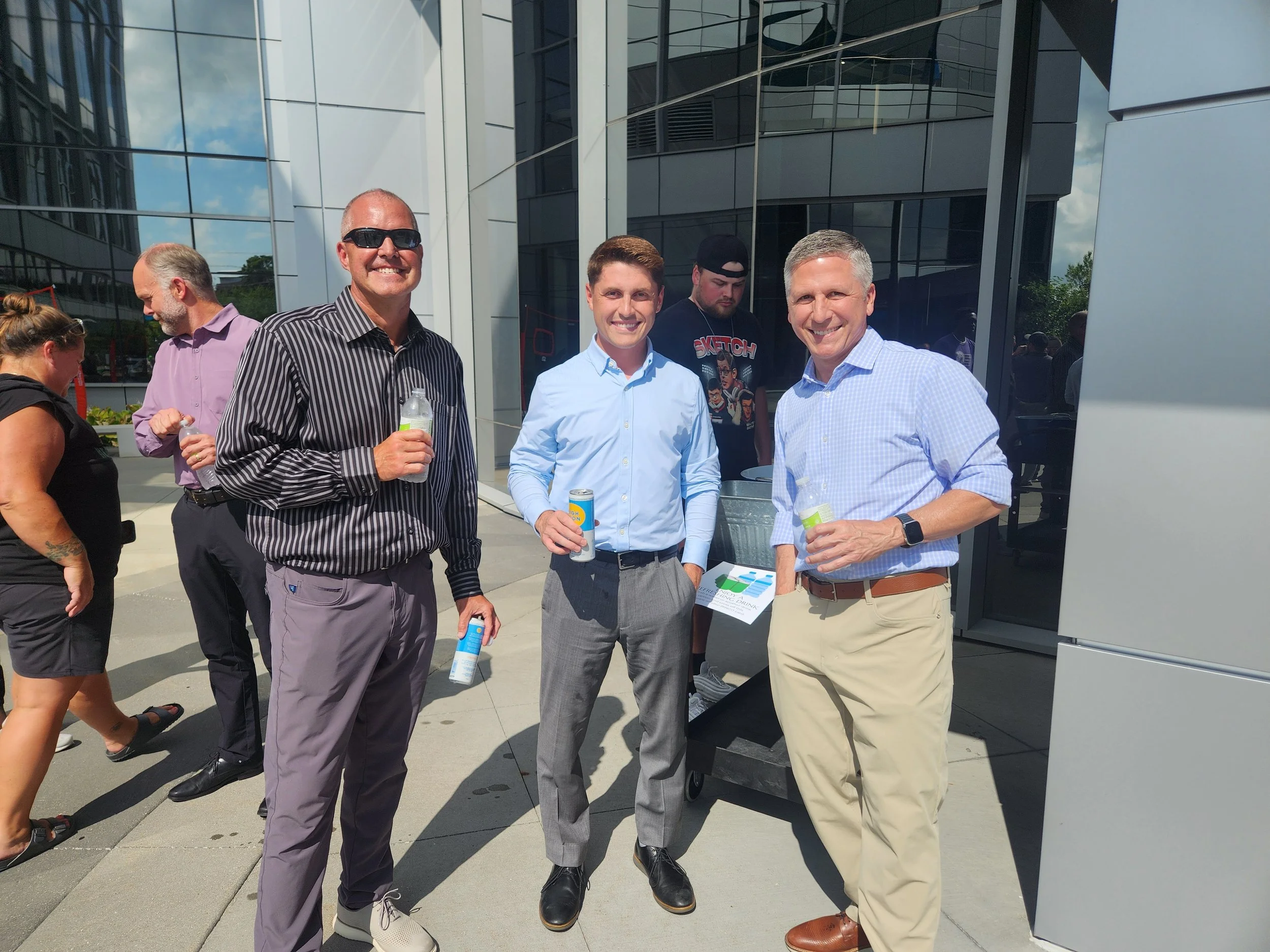 Three men standing outside a modern glass building, smiling and holding drinks, with other people in the background.