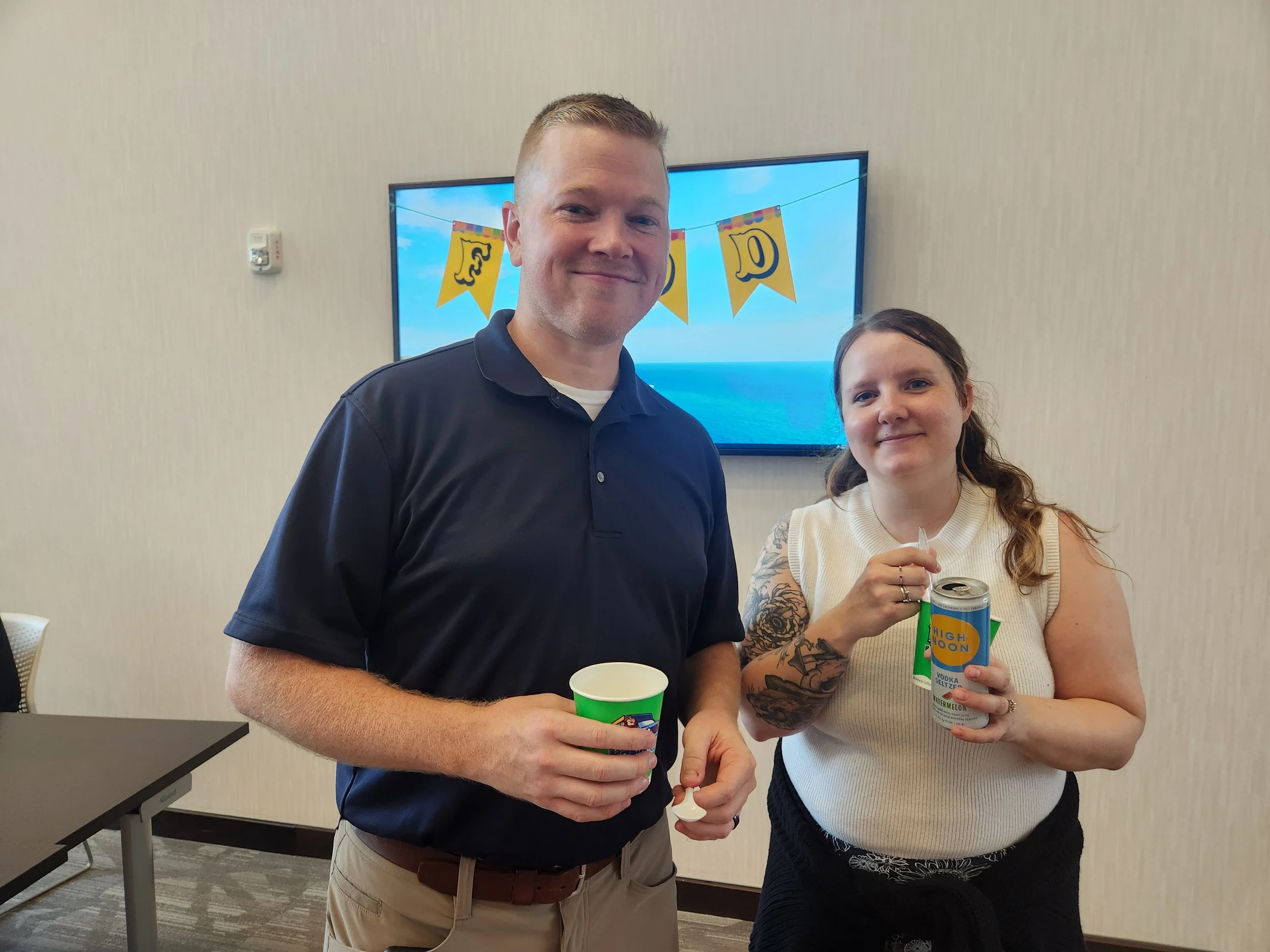 A man and a woman standing indoors, both smiling and holding drinks at a celebration. The man is wearing a dark polo shirt, and the woman has tattoos on her arm and is wearing a cream-colored sleeveless top. Behind them is a screen with a blue ocean 