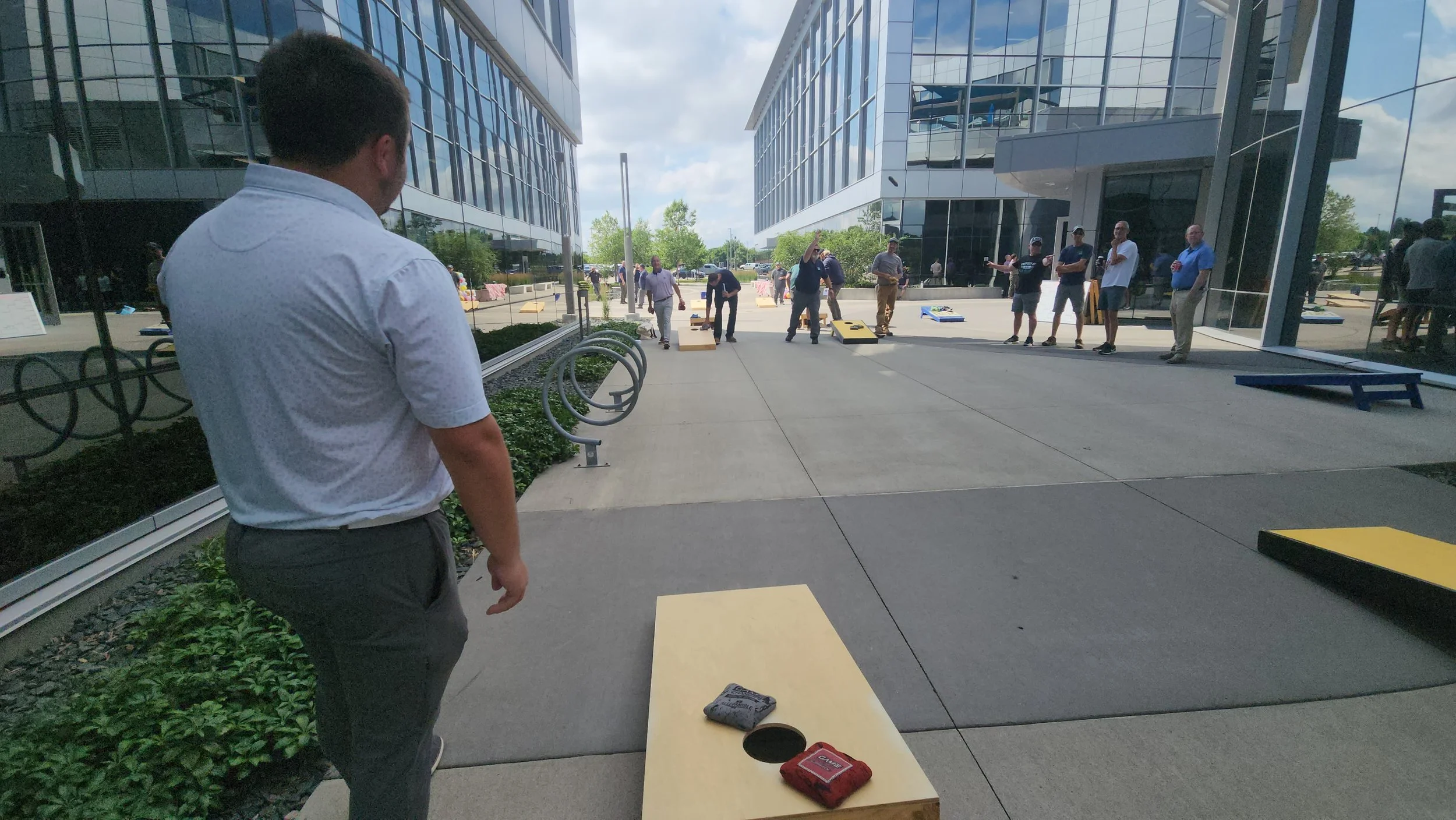 People playing cornhole outside of a modern office building with glass walls, some watching and others participating in the game.