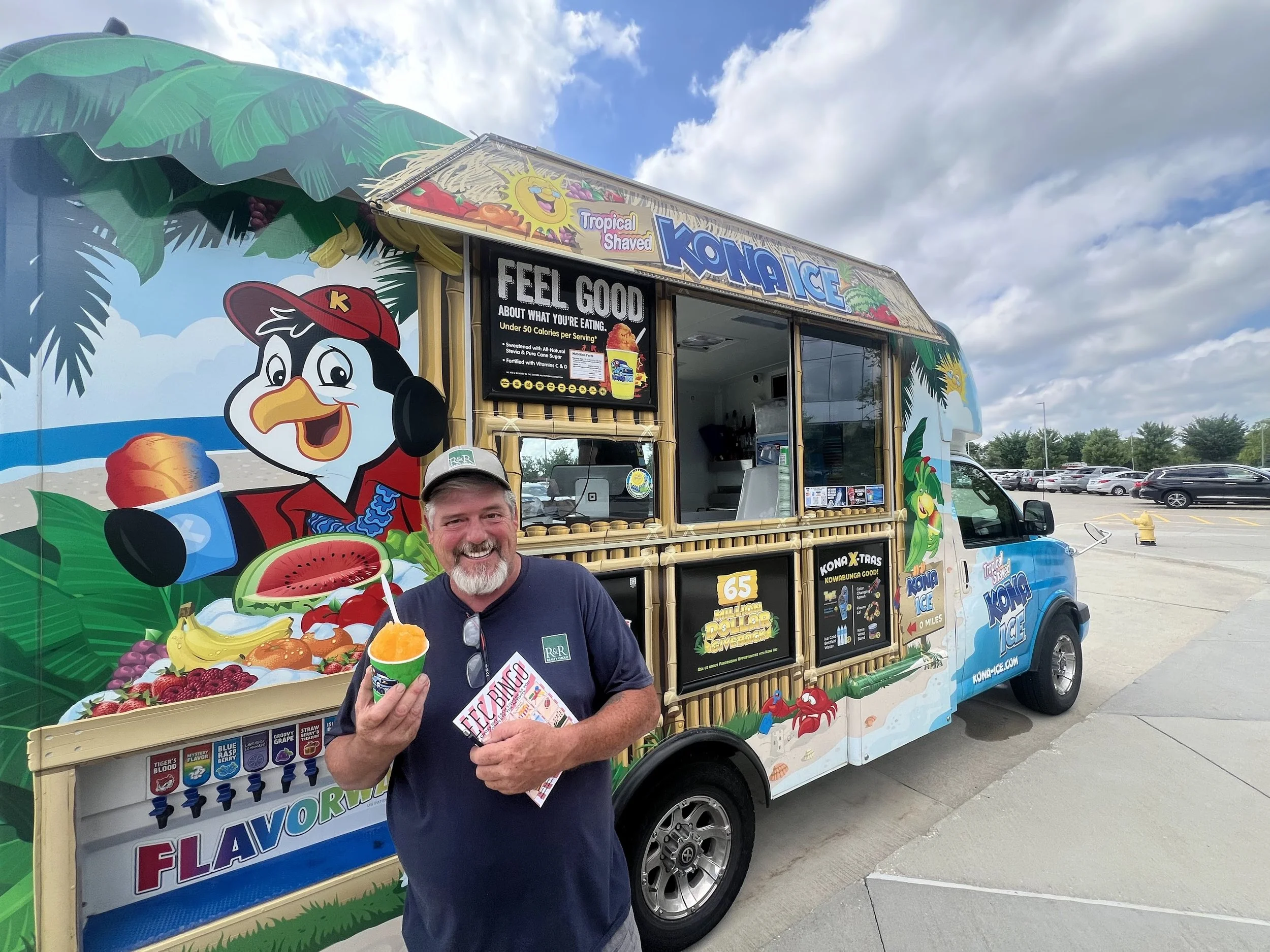 Smiling man holding a frozen treat in front of a colorful Kona Ice truck with tropical-themed artwork and signage.