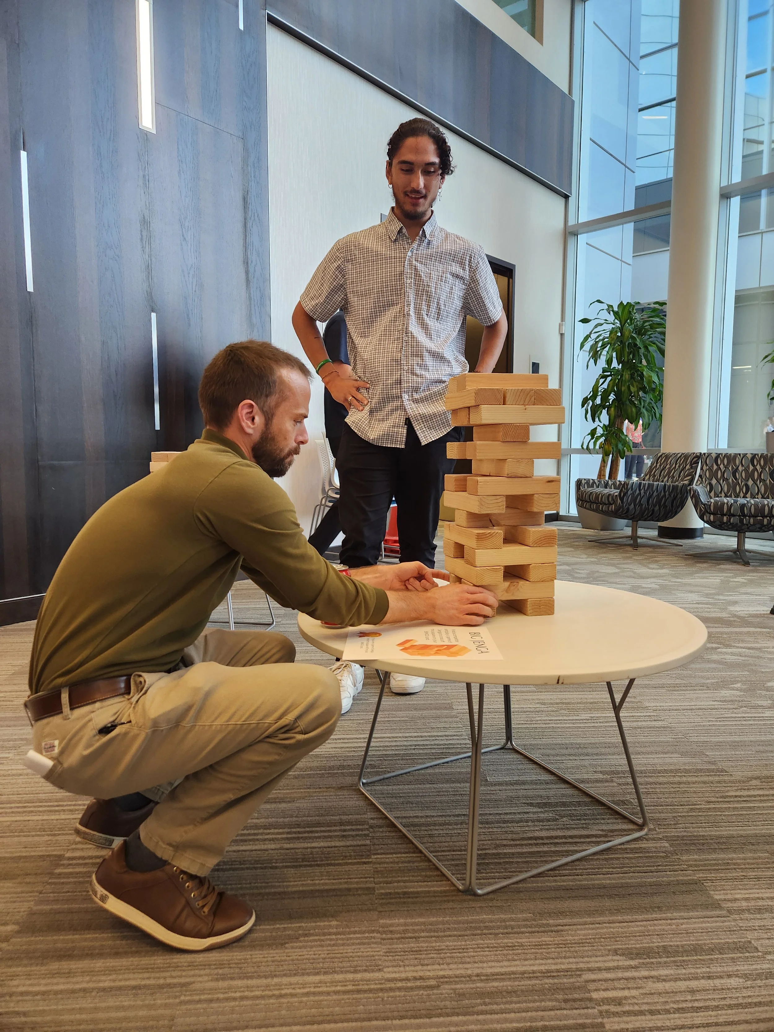 Two men playing a large outdoor Jenga game in a modern office lobby.