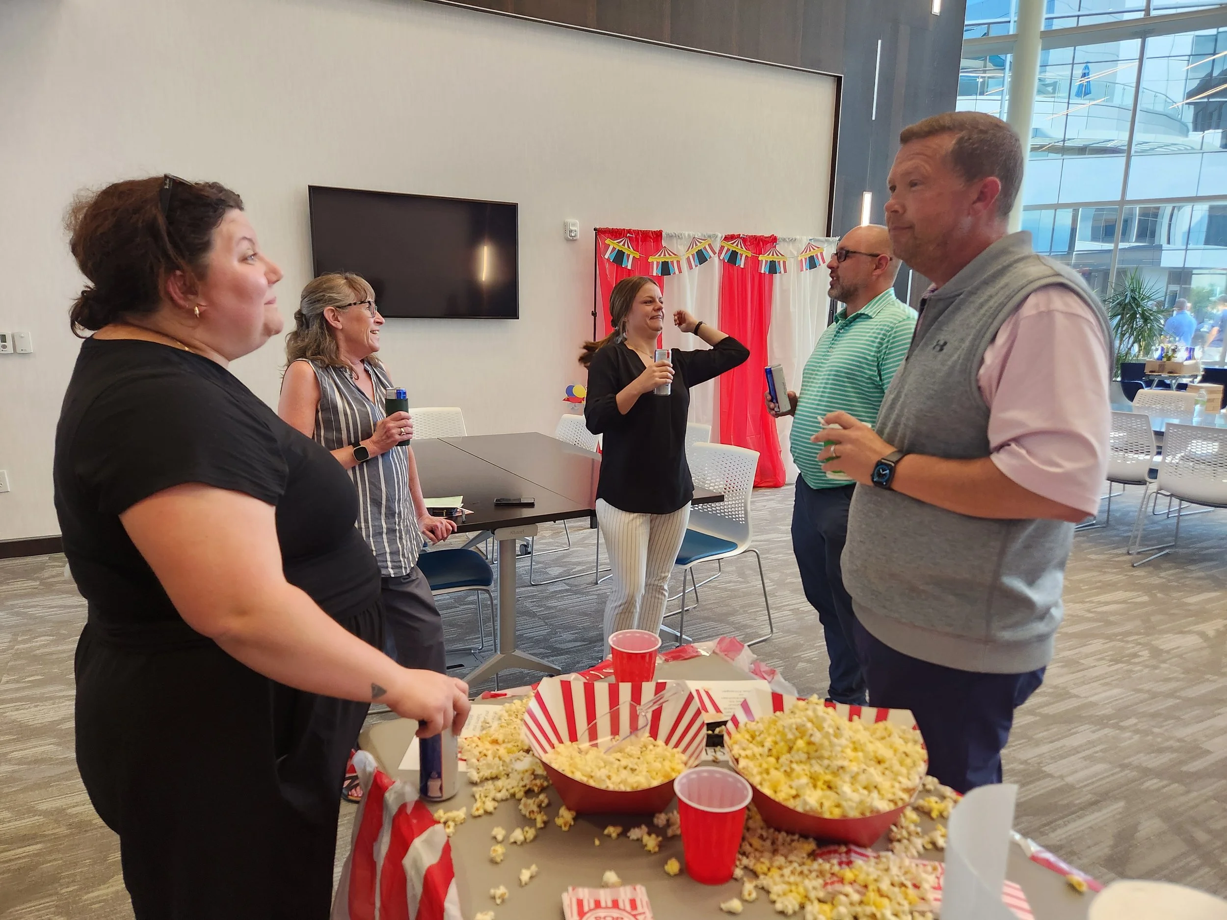 Group of five people standing and talking around a table with popcorn and red cups during a celebration with patriotic decorations.