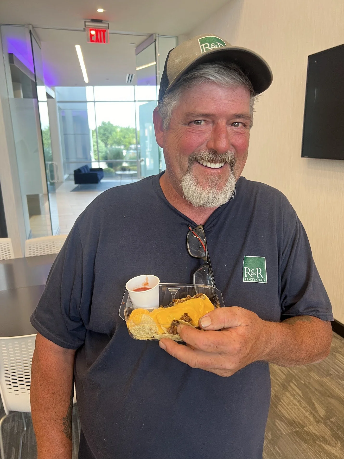 A smiling man with gray hair and beard wearing a R&R Realty Group shirt and cap, holding a plastic tray with nachos and a small container of salsa inside a modern office lobby.