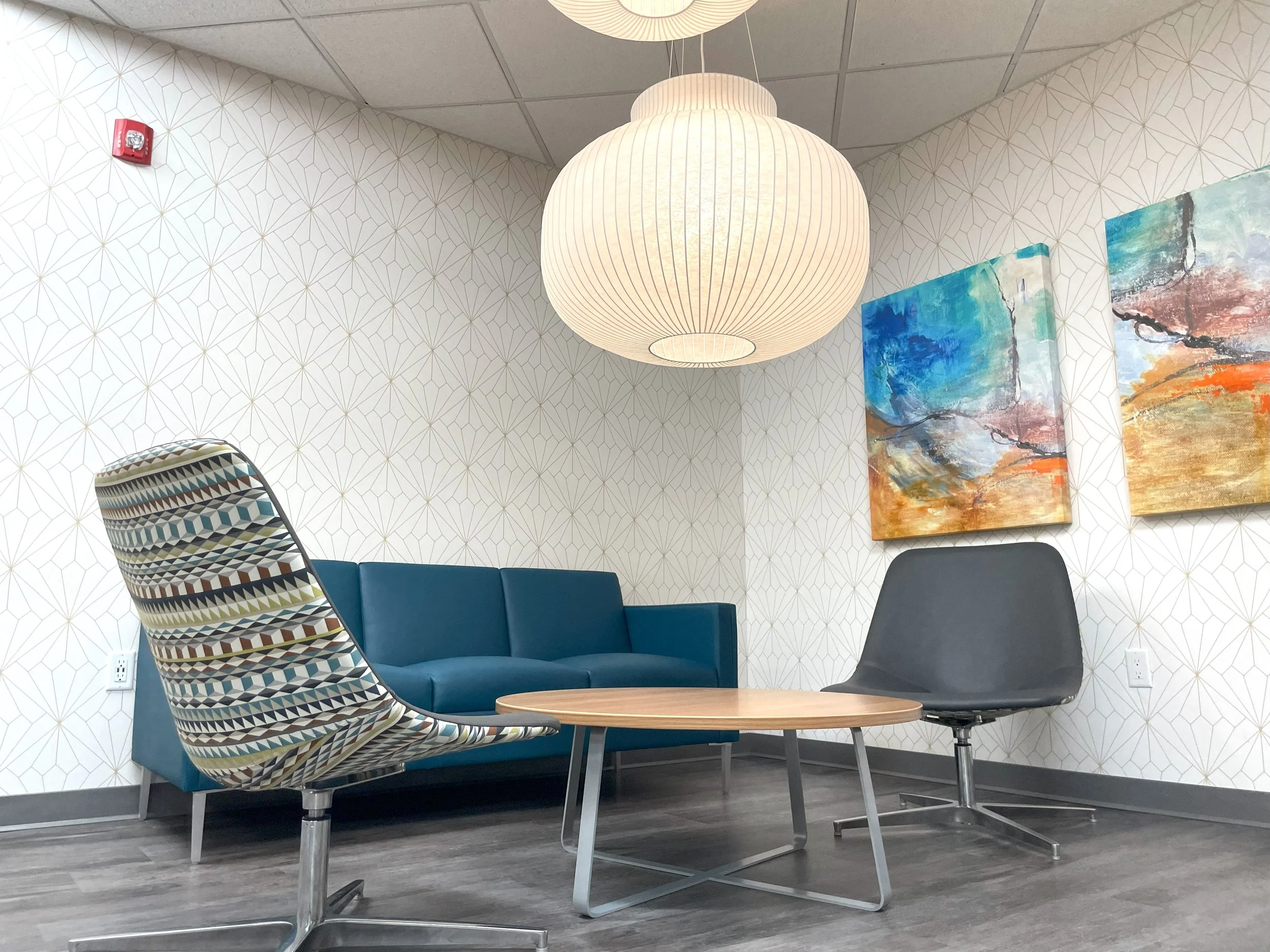 A modern waiting area with a blue sofa, a patterned armchair, a black swivel chair, a round wooden coffee table, abstract paintings on the wall, and a large white paper lantern hanging from the ceiling.