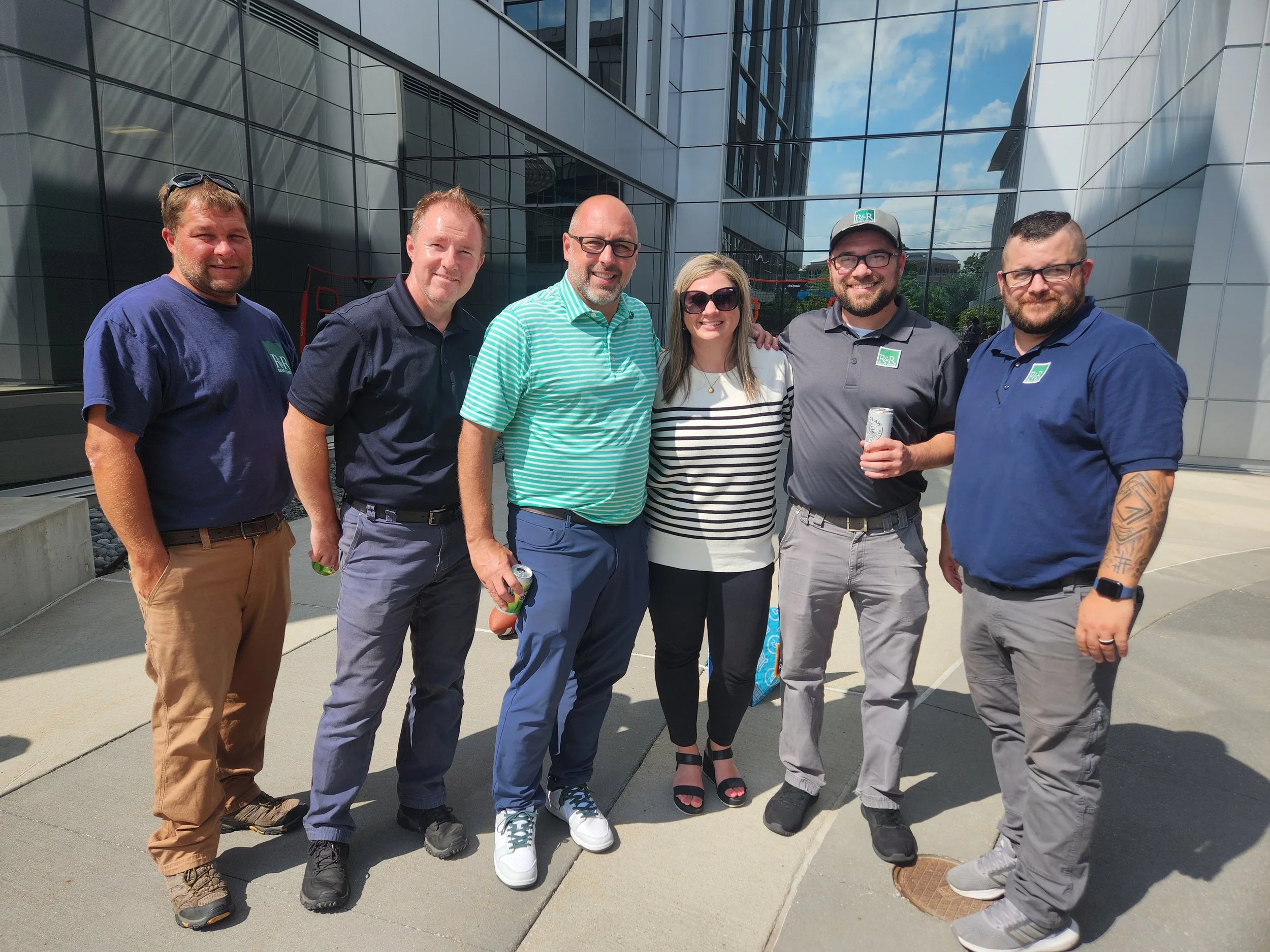 Group of six people, five men and one woman, standing outside in front of modern office buildings, smiling for the camera on a sunny day.