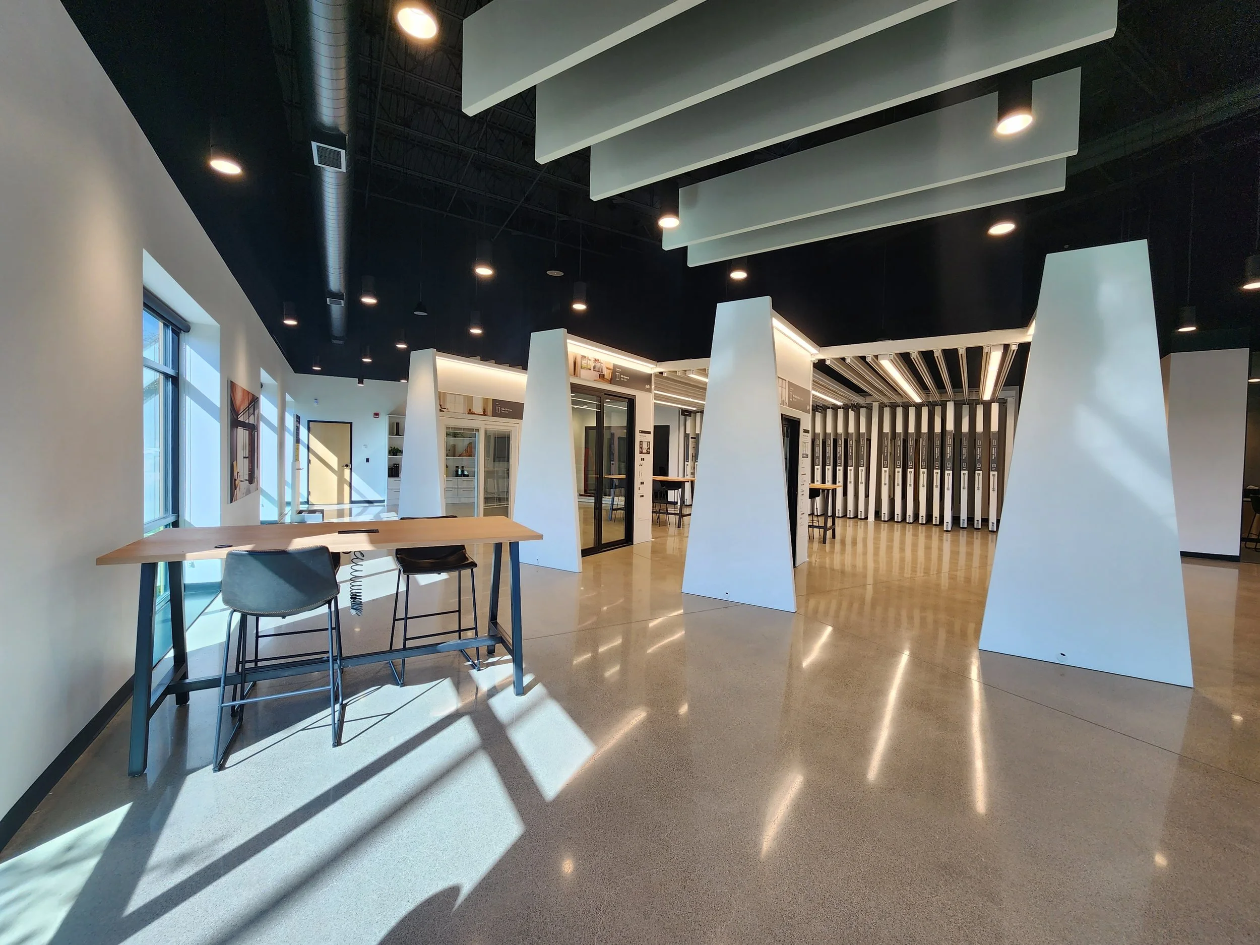 Modern office space with high ceilings, large windows, a wooden table with two black chairs, and white structural beams.